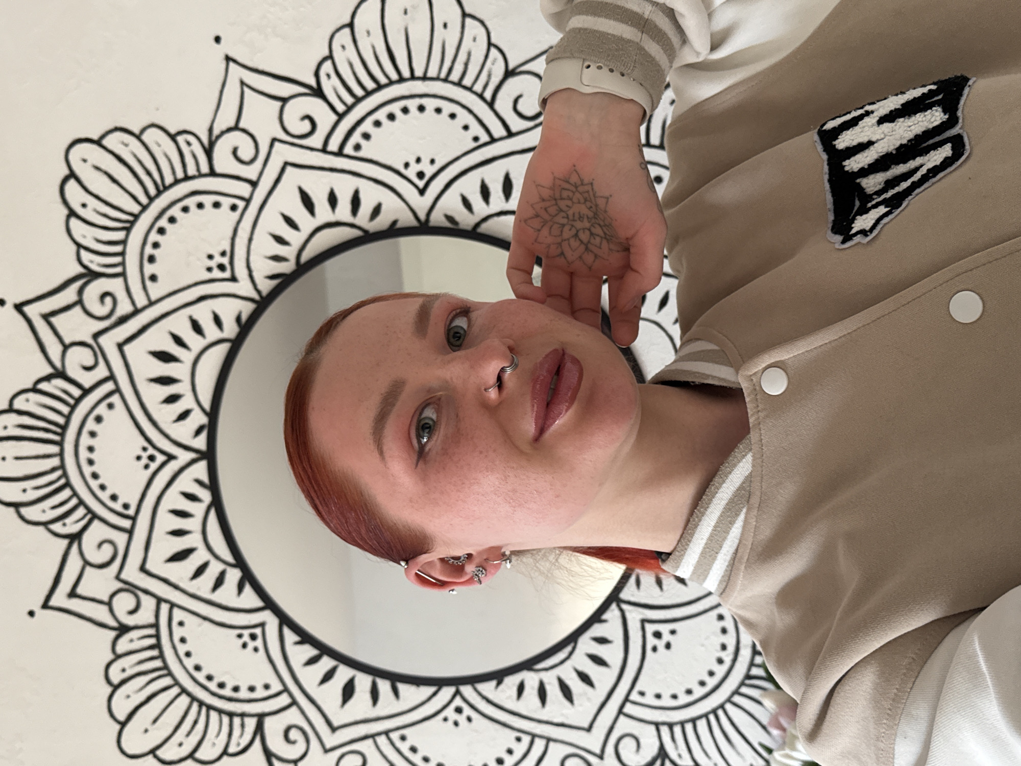 The image, rotated 90 degrees clockwise, features a young person with fair skin, freckles, and reddish hair, looking directly at the camera with a neutral expression. She has light-colored eyes and prominent facial and ear piercings, including a nose ring, a septum ring, an industrial bar, and multiple helix, tragus, and lobe piercings.
She is wearing a beige or tan bomber-style jacket with white and striped cuffs, covering a white inner garment. On the left side of her jacket, a black and white patch resembling a stylized letter, possibly "S" or "B", with a zebra stripe pattern is visible. White snap buttons run down the front of the jacket. Her left hand is raised, showcasing a detailed mandala-style tattoo on her palm that includes the words "ARTE ART".
The background consists of a white wall adorned with a large, intricate black and white mandala pattern. A circular mirror is positioned within this pattern, partially reflecting a plain white surface. The overall setting appears to be indoors, possibly a room decorated with this unique wall art.