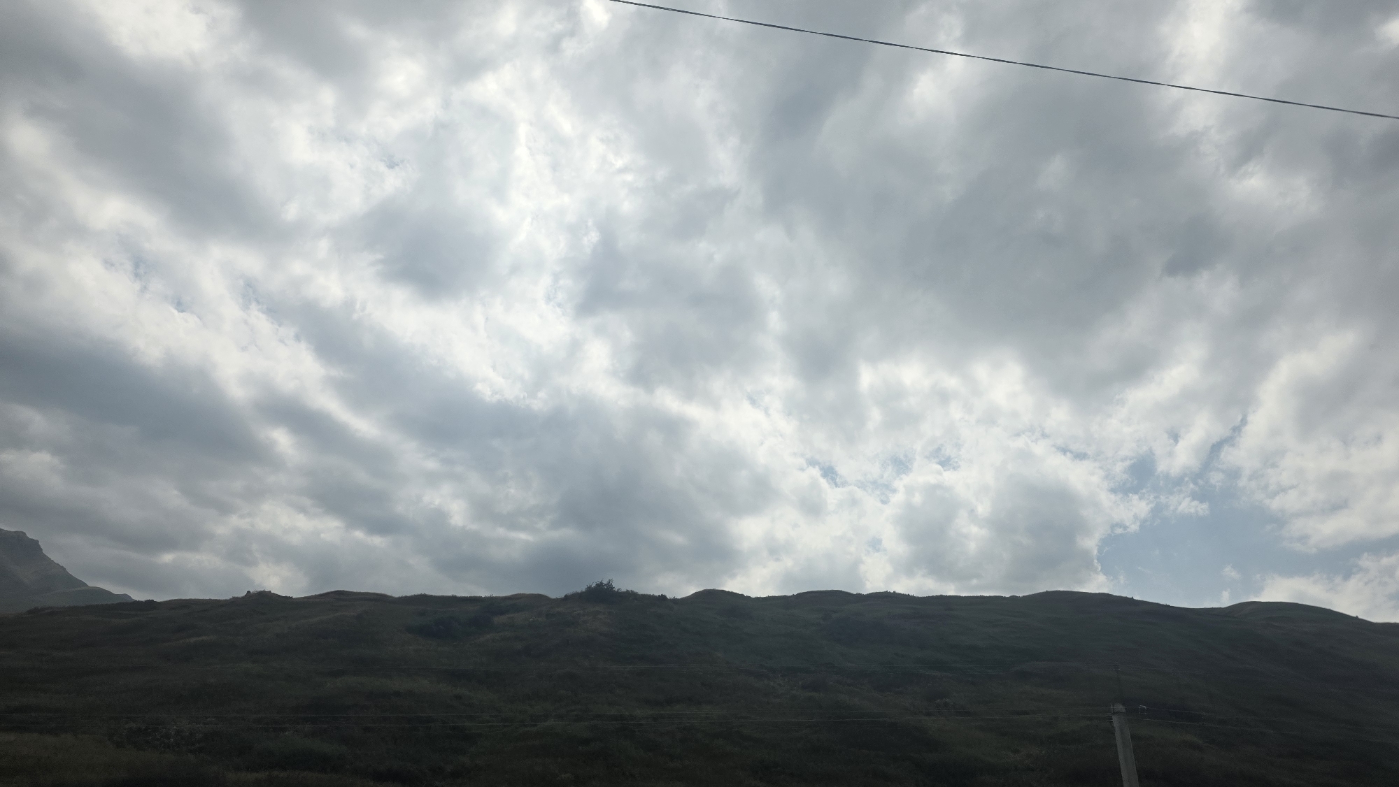 The image shows a dark, undulating landscape under a cloudy sky. The foreground is a dark, hilly terrain covered in vegetation, appearing as a silhouette against the brighter sky. The sky is filled with dense, grey and white clouds, with patches of lighter blue sky visible in between. A thin, dark horizontal line, possibly a wire or cable, stretches across the top portion of the image. The scene appears to be a natural landscape, possibly in a rural area, and the lighting suggests it could be late afternoon or early morning, or a day with overcast weather. There are no visible people or specific man-made structures in the immediate foreground or on the hills, though there might be very faint lines suggesting fences or power lines in the distance. The overall impression is one of a vast, natural expanse under a dramatic sky.