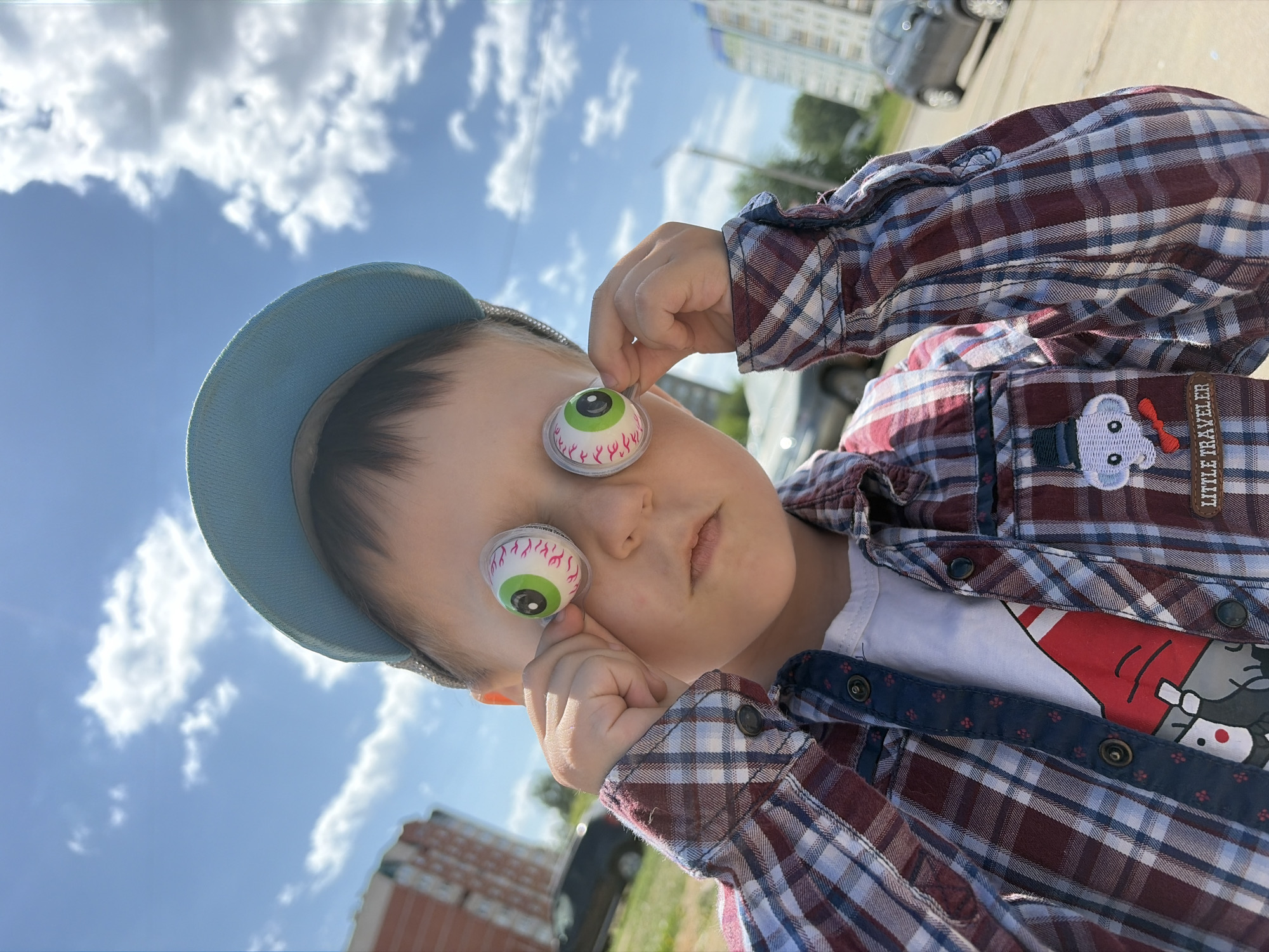 A close-up shows a child standing outside on a sunny day in Chaikovsky, Russia. The child is wearing a light blue baseball cap and a plaid shirt with an embroidered teddy bear and the words "Little Traveler" on the chest. The child is holding two eyeball candies in front of their eyes. The background features a blue sky with scattered clouds, buildings, parked cars, and green grass.