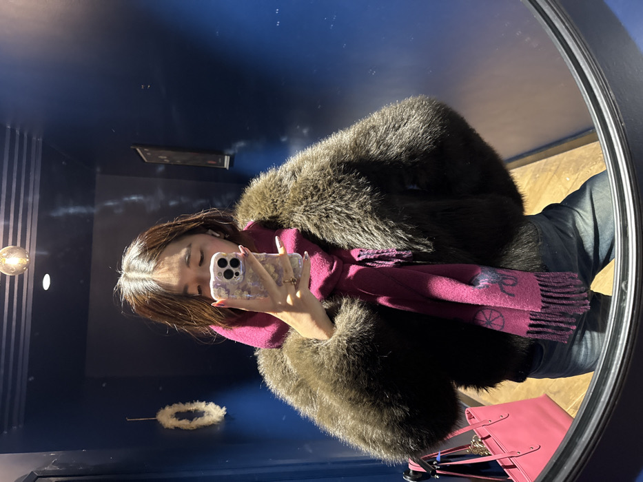 The image displays a mirror selfie of a person taken indoors within what appears to be a small room or hallway. The person, an individual with medium-length brown hair, wears a dark brown faux fur or fur coat, dark blue jeans, and a magenta-colored fringed scarf featuring a dark blue or purple equestrian logo resembling the Ralph Lauren polo player. A white wireless earbud is visible in their right ear, and a ring is present on their right ring finger. They hold a smartphone with a triple camera system, encased in a light-colored, patterned case, obscuring the upper portion of their face. To the left of the person's reflection, a hot pink leather tote bag with gold-colored hardware hangs on a black stand. The interior environment features dark blue walls, a light brown wooden floor, and an overhead exposed filament light bulb. A white, textured wreath-like object hangs on the left wall, and a dark-framed picture or artwork is on the right wall. The exact location within Paris, France, cannot be determined from the visual information provided in the image.