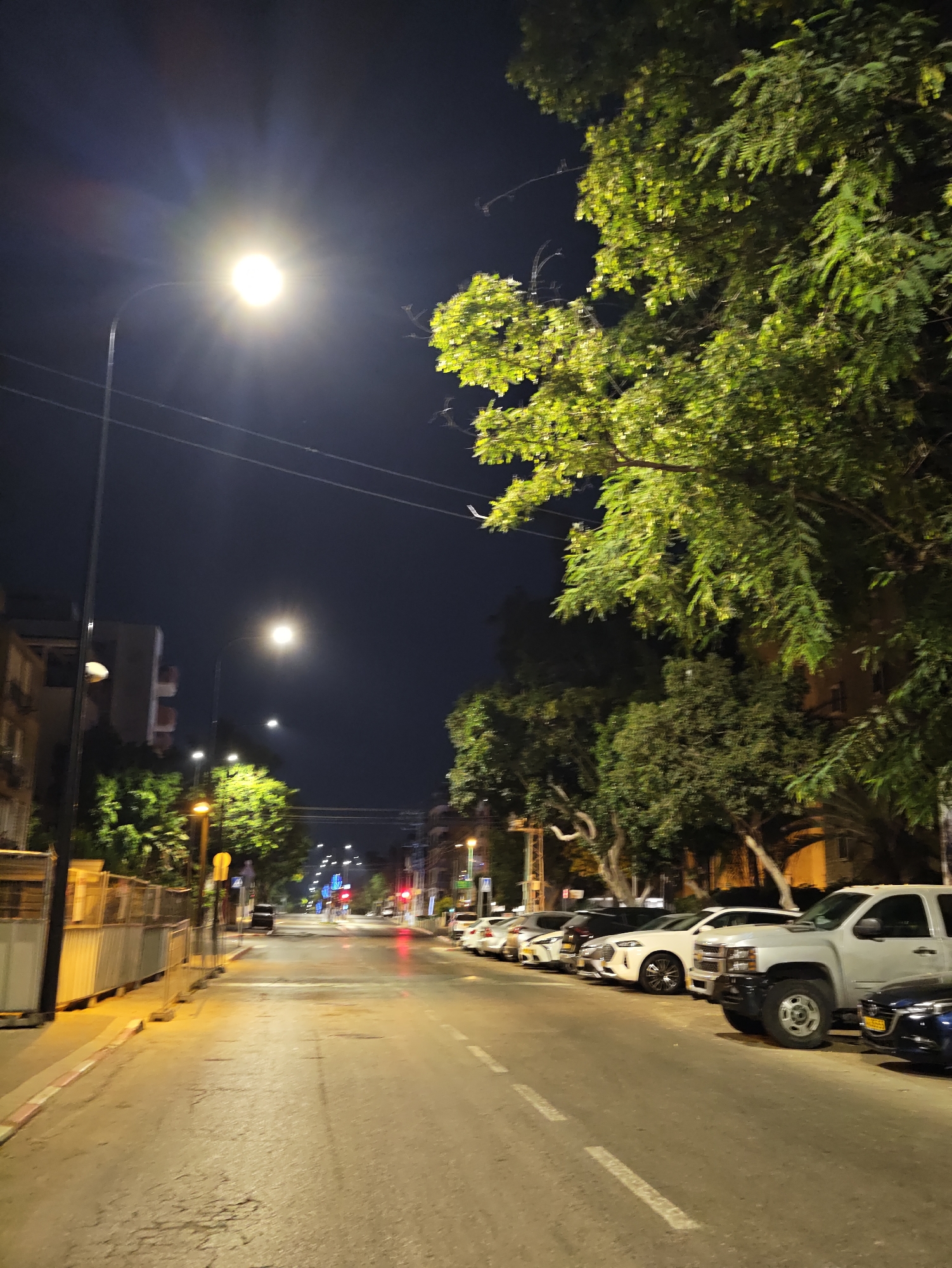 The image depicts a street scene at night in Petah Tikva, Israel. The view is from a street level perspective, looking down an empty street lined with parked cars and trees. A bright streetlight illuminates the scene from the left, casting long shadows and creating lens flare. In the foreground, a row of parked vehicles is visible, including a silver pickup truck in the right foreground. The street surface appears to be asphalt, with some cracks and wear. On the left side of the street, a construction barrier is present, and further down, apartment buildings are visible. The right side of the street is dominated by dense foliage from trees. Traffic lights are visible in the distance, with one showing a red light. The sky is dark and clear.
