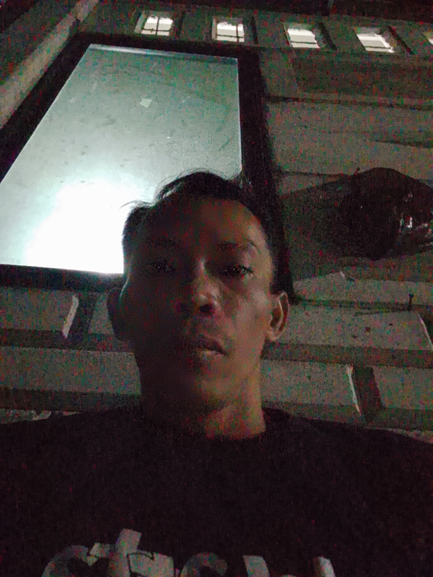A dimly lit, low-angle selfie shows a person with short, dark hair and a dark t-shirt. The person faces the camera with a neutral expression. Behind the person, a structure with small windows is visible. The location is Bukit Ketok, Indonesia.