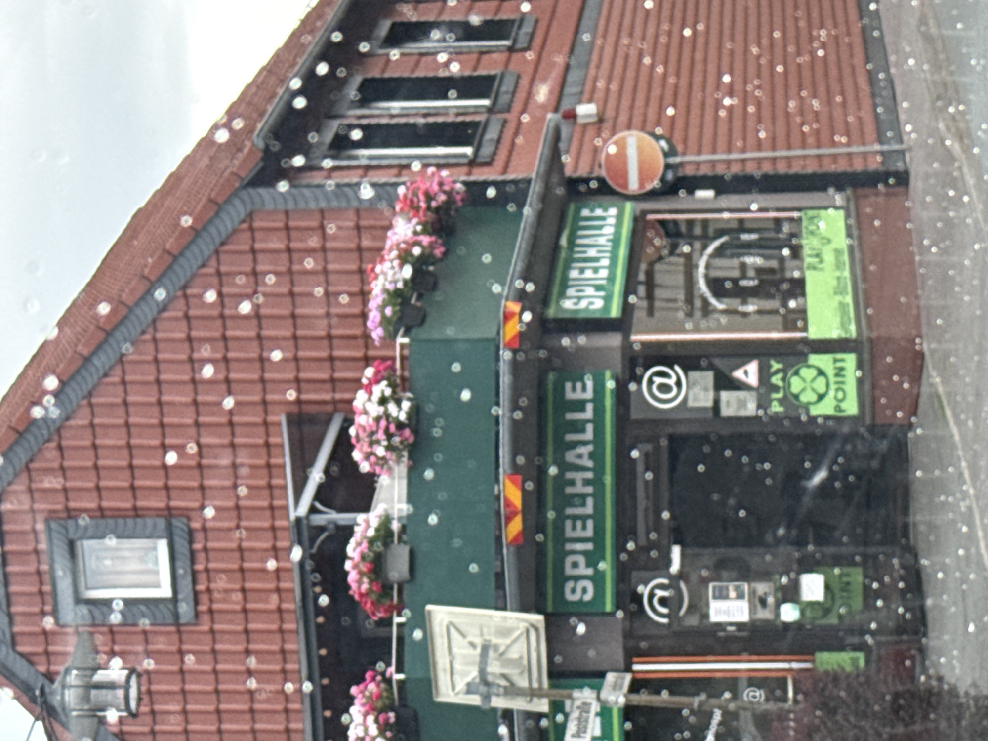 The image shows a view of the facade of a building, likely a commercial establishment, with the German text "SPIELHALLE" visible on a green sign, indicating it is an arcade or gaming hall. The building has a red tiled roof and brick walls. There are flower boxes with pink and white flowers on a balcony. The scene is captured through a window, as evidenced by numerous circular water droplets scattered across the image. A street sign with "Poststraße" is partially visible. A red and white circular traffic sign, likely a "No Entry" sign, is present to the right of the entrance. The sky is overcast. The image is undated and the time of day is indeterminate due to the lighting conditions. The general setting appears to be a street in a German town, with the specific location identified as Bad Sachsa, Germany. No individuals are visible in the frame.