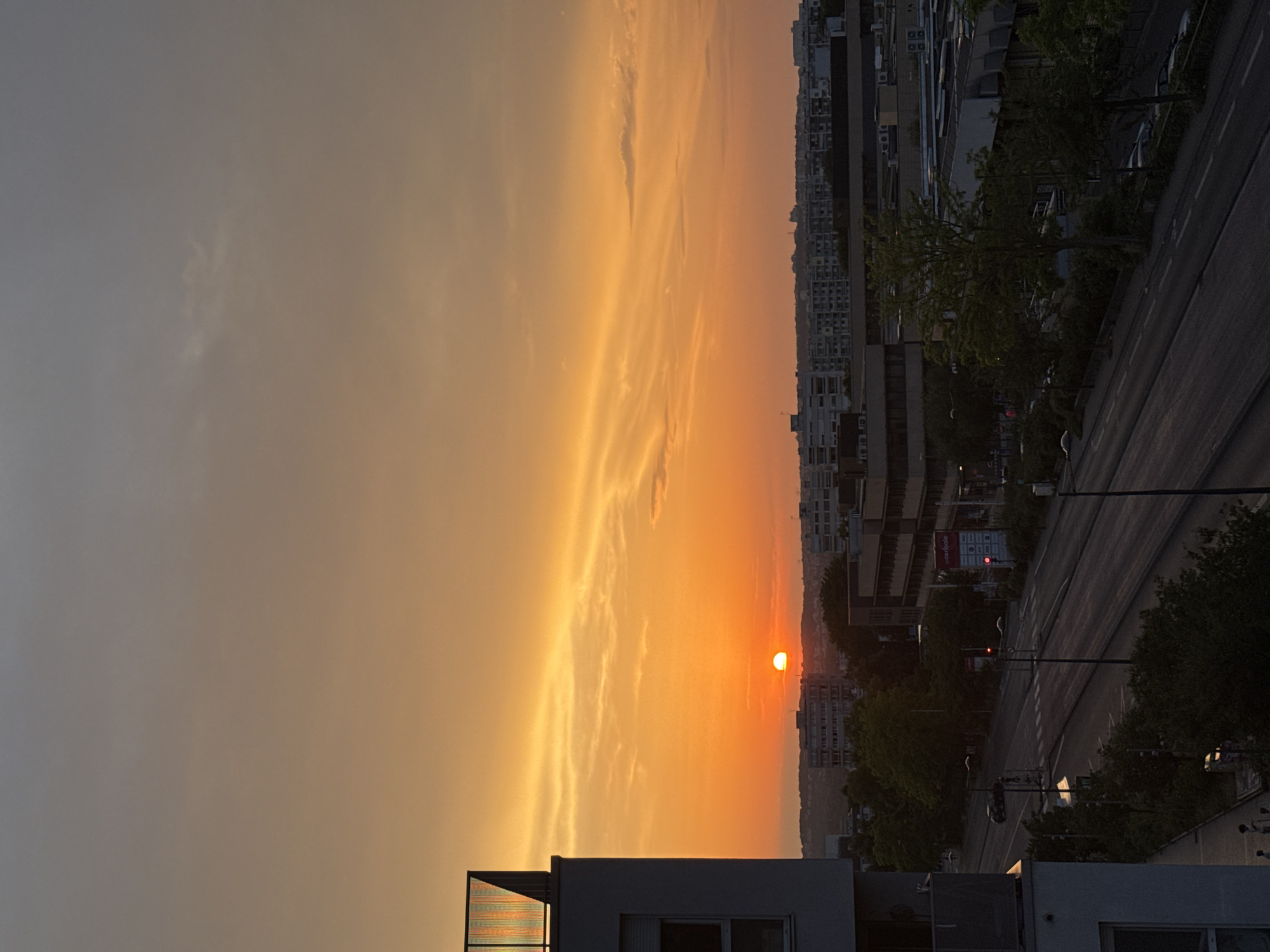 The image depicts a sunset over a city skyline. The sun, a bright orange orb, is partially obscured by the horizon, casting a warm glow across the sky. Wispy clouds stretch across the upper portion of the image, tinged with shades of orange and yellow. In the foreground, the edge of a modern building is visible on the left, with a balcony railing at the top. Below the building, a street runs horizontally, with a few cars and streetlights visible. The middle ground features a line of buildings, including some apartment complexes and commercial structures. Trees and greenery are interspersed throughout the scene, softening the urban landscape. The overall impression is one of a peaceful evening in a city.