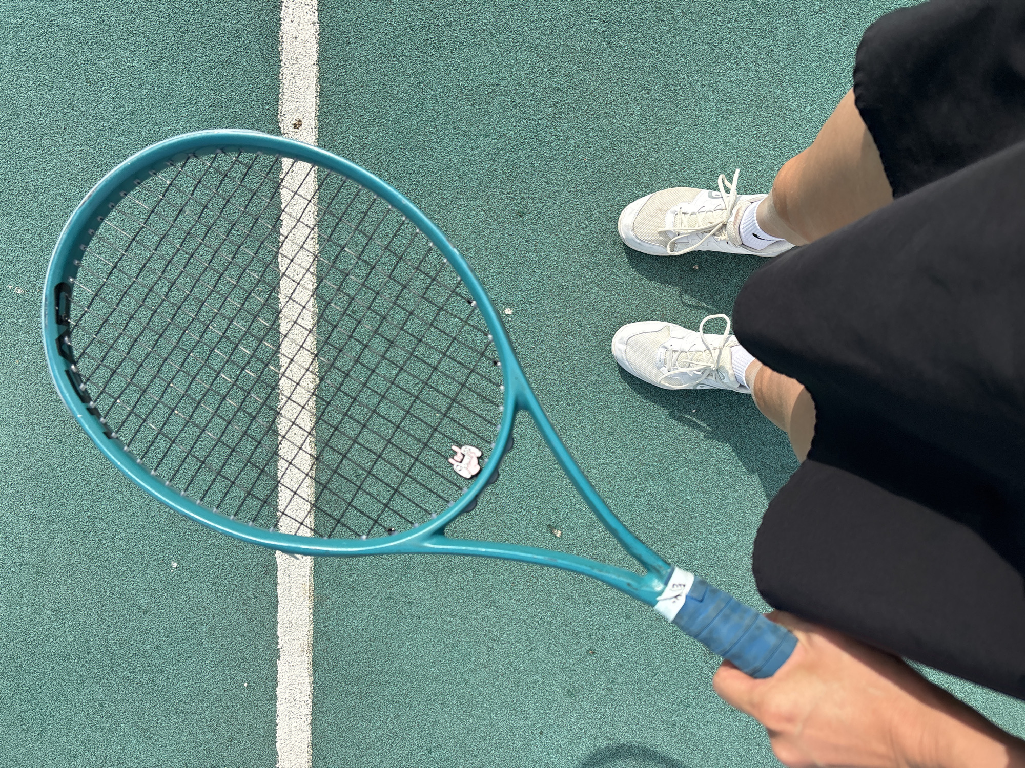 A person stands on a teal-colored tennis court with white lines, wearing white athletic shoes and black shorts. They hold a light blue tennis racket with a white object attached to the strings. The location of Chaikovsky, Russia, cannot be confirmed from the image.