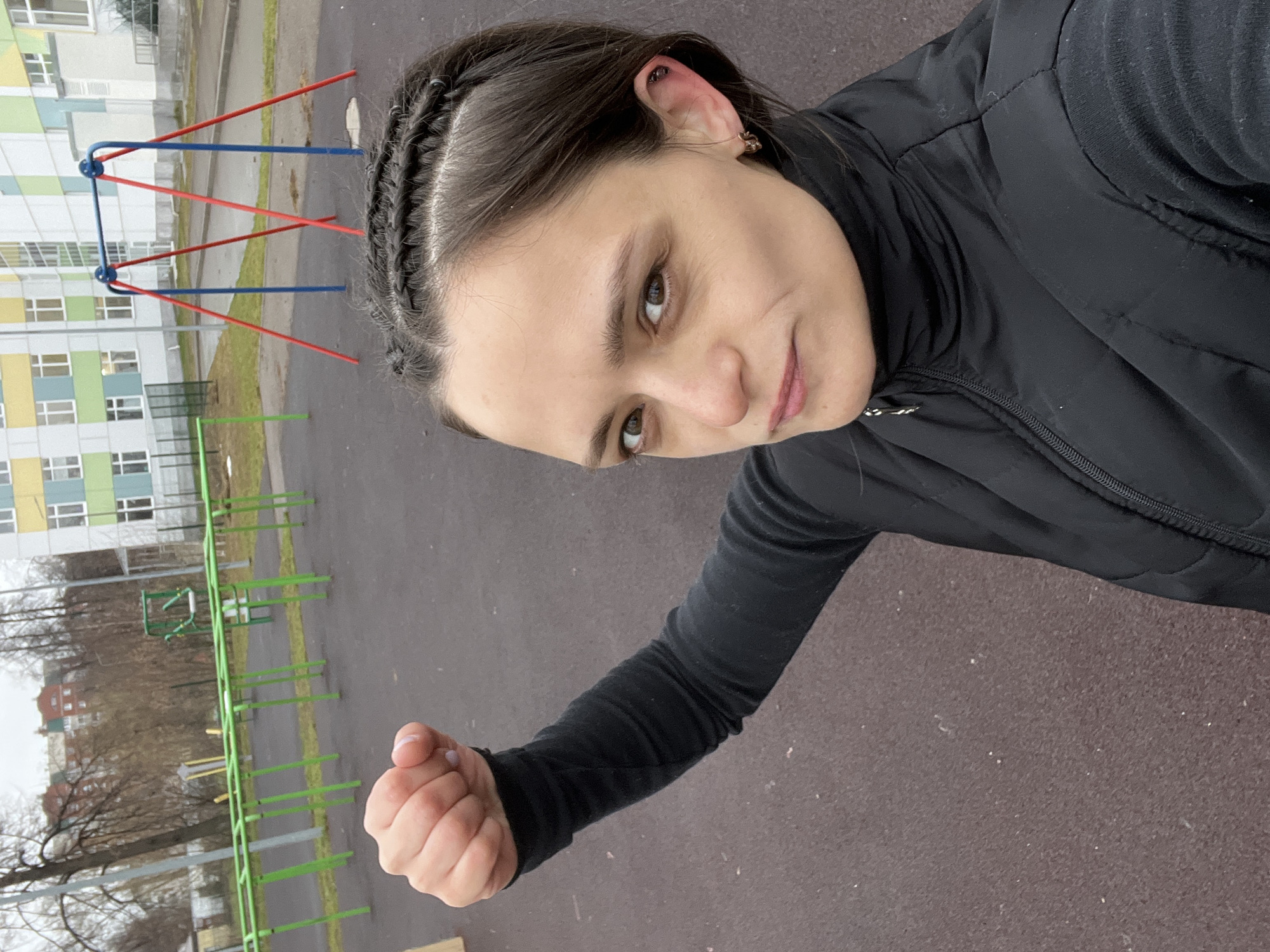 A young woman is taking a selfie outdoors. She has dark hair braided on the sides and is wearing a black puffer vest and a black long-sleeved shirt. She is holding her fist up in front of the camera. In the background, there is a playground with red, blue, and green play equipment, a building with light green and yellow sections, and bare trees. The location is Chaikovsky, Russia.