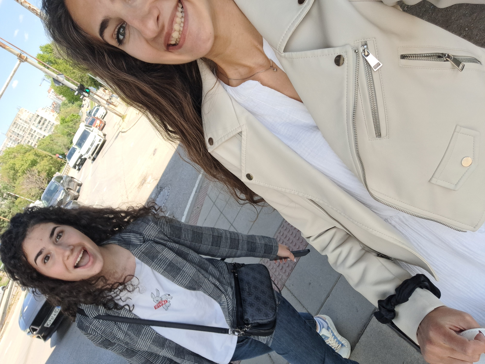 Two young women are taking a selfie outdoors on a sidewalk or pedestrian area in Sofia, Bulgaria. The woman in the upper part of the frame has long, dark hair and is wearing a light-colored leather jacket over a white top. She is smiling and looking directly at the camera. The other woman is wearing a gray plaid blazer over a white T-shirt with a cartoon bunny, blue jeans, and sneakers. She has curly dark hair and is also smiling broadly. She has a black purse slung across her body. In the background, there is street traffic, buildings, and a traffic light pole. The pavement is made of rectangular gray stones.