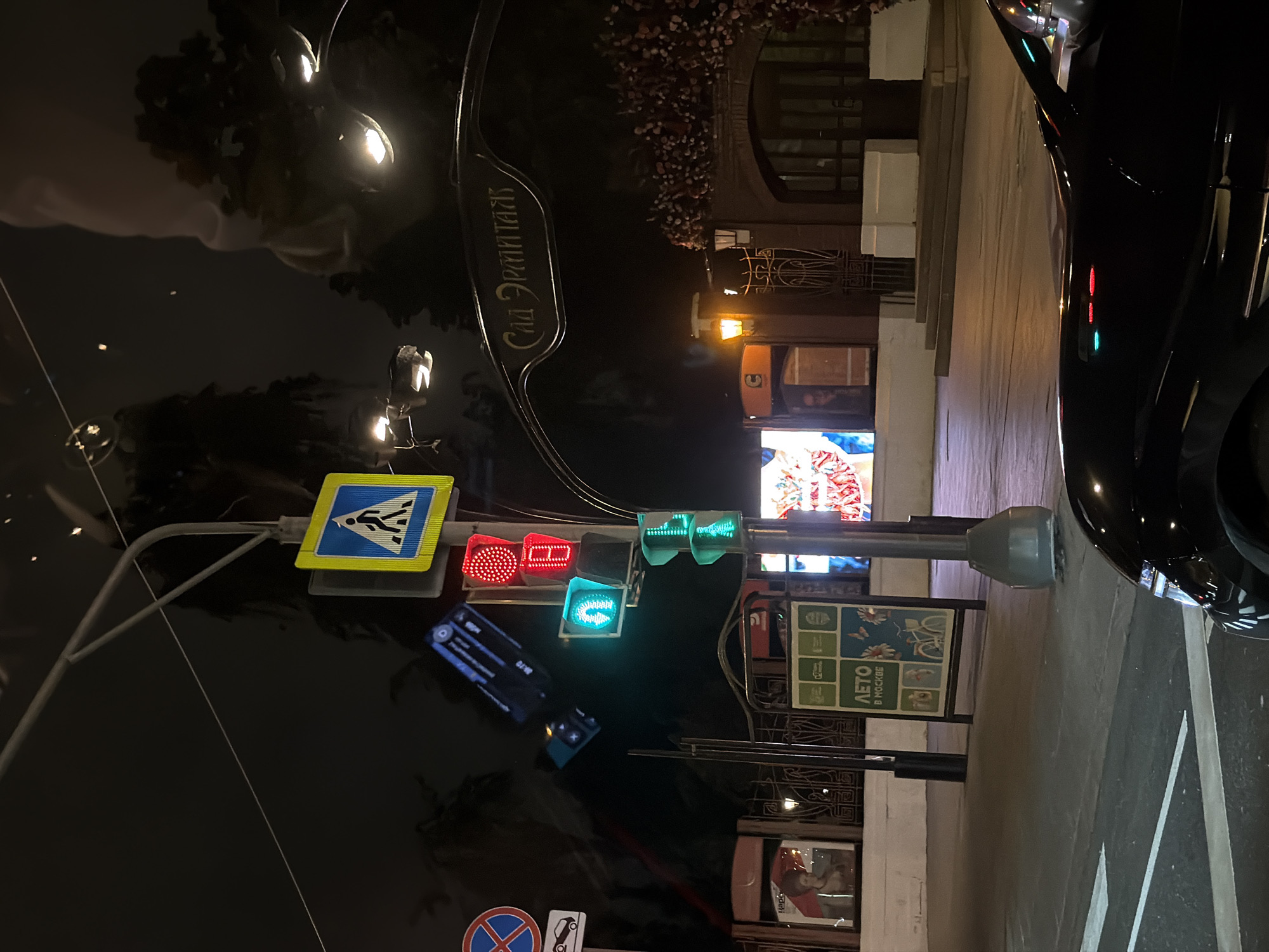 Captured at night in Moscow, Russia, the image features a street scene reflected in a car's hood. A street sign for "Sad Ermitazh" (Hermitage Garden) is visible above the reflection. A pedestrian crossing sign is near a traffic signal indicating both red and green lights are active. A smaller light below the traffic signal is green. A sign advertising "Leto v Moskve" (Summer in Moscow) is prominently displayed. The surrounding environment includes buildings, signage, and street infrastructure. The presence of a black car, the illuminated signs and traffic lights, and the streetscape contribute to an urban nighttime environment.