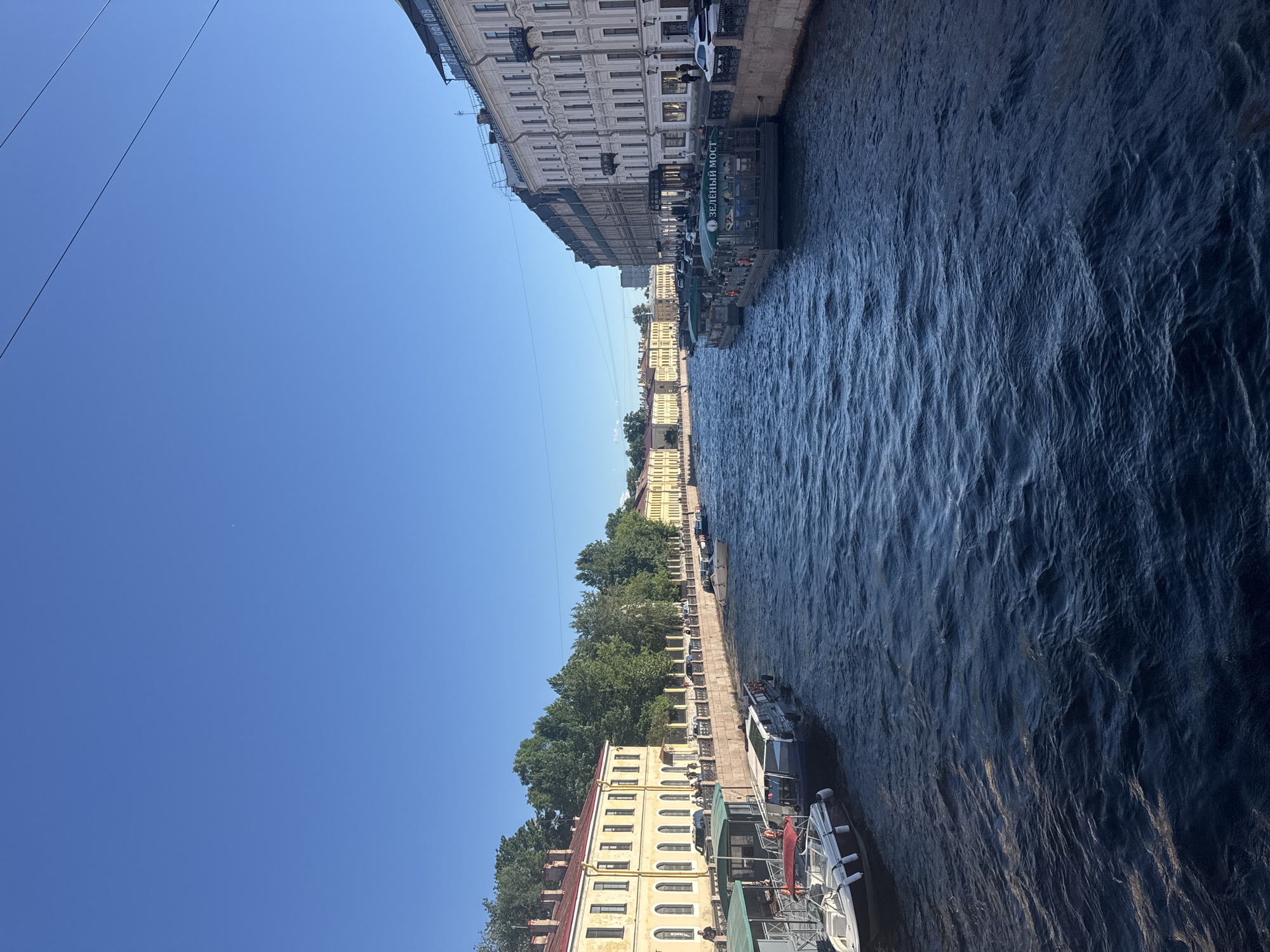 This media file is a vertical, eye-level shot of a canal in Saint Petersburg, Russia, on a clear, sunny day. The canal is dark blue with rippling water, suggesting a moderate breeze. Along the left bank of the canal, a row of historic buildings with light-colored facades and arched windows are visible. The buildings are four to five stories tall and appear to be from an older architectural period.
Several boats are docked along the canal's edge. One prominent boat has green awnings and the visible text "ЗЕЛЕНЫЙ МОСТ" (Zelenyy Most, meaning "Green Bridge" in Russian). Other boats, including white passenger vessels, are also present. Trees line the bank behind the buildings, and some greenery is visible on the boats themselves. Power lines stretch across the clear blue sky. The overall scene depicts a serene urban canal, likely a popular spot for boat tours or leisurely strolls. The bright sunlight and clear sky indicate it is daytime, possibly mid-morning or afternoon, with pleasant weather. There are no discernible people in the foreground, but the presence of docked boats suggests activity may be occurring or has recently occurred.
