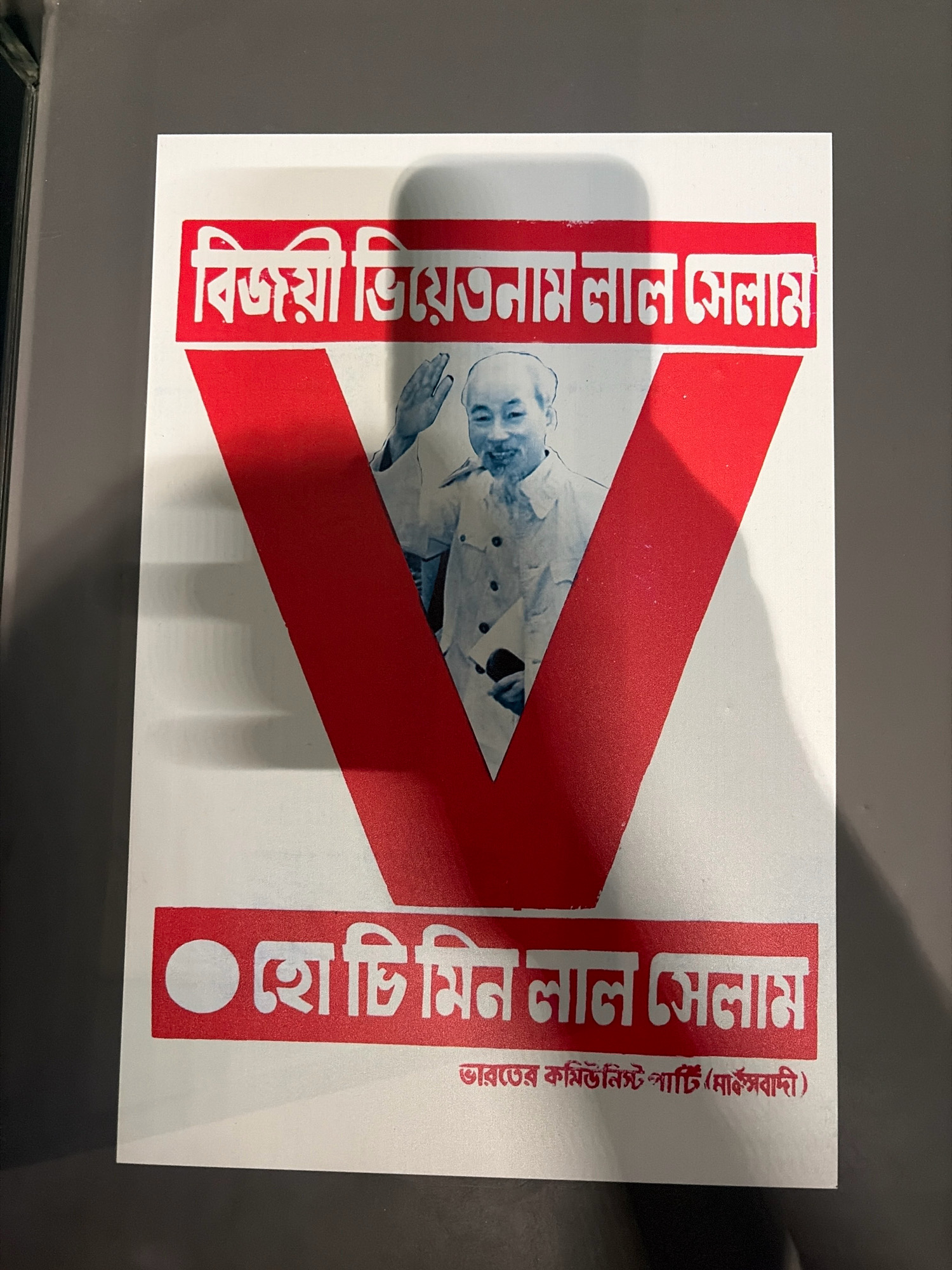The image shows a poster with a large red "V" shape as a prominent design element. Inside the V, there is a black and white photograph of a man with a mustache and short hair, wearing a light-colored shirt, waving. The man appears to be of East Asian descent and is smiling.
Above the "V" and the photograph, there is a red banner with white text in Bengali script. The text reads "বিজয়ী ভিয়েতনাম লাল সেলাম" which translates to "Victorious Vietnam Red Salute."
Below the "V" and the photograph, there is another red banner with a white circle on the left and white text in Bengali script to its right. This text reads "হো চি মিন লাল সেলাম" which translates to "Ho Chi Minh Red Salute."
Beneath this banner, there is a line of smaller red text in Bengali script that says "ভারতের কমিউনিস্ট পার্টি (মার্কসবাদী)" which translates to "Communist Party of India (Marxist)."
The poster is set against a white background and appears to be a political or commemorative item. The image does not contain any identifiable landmarks or elements that would place it specifically in the city of Đà Nẵng, Vietnam. The content of the poster suggests a connection to Vietnam and the Communist Party.