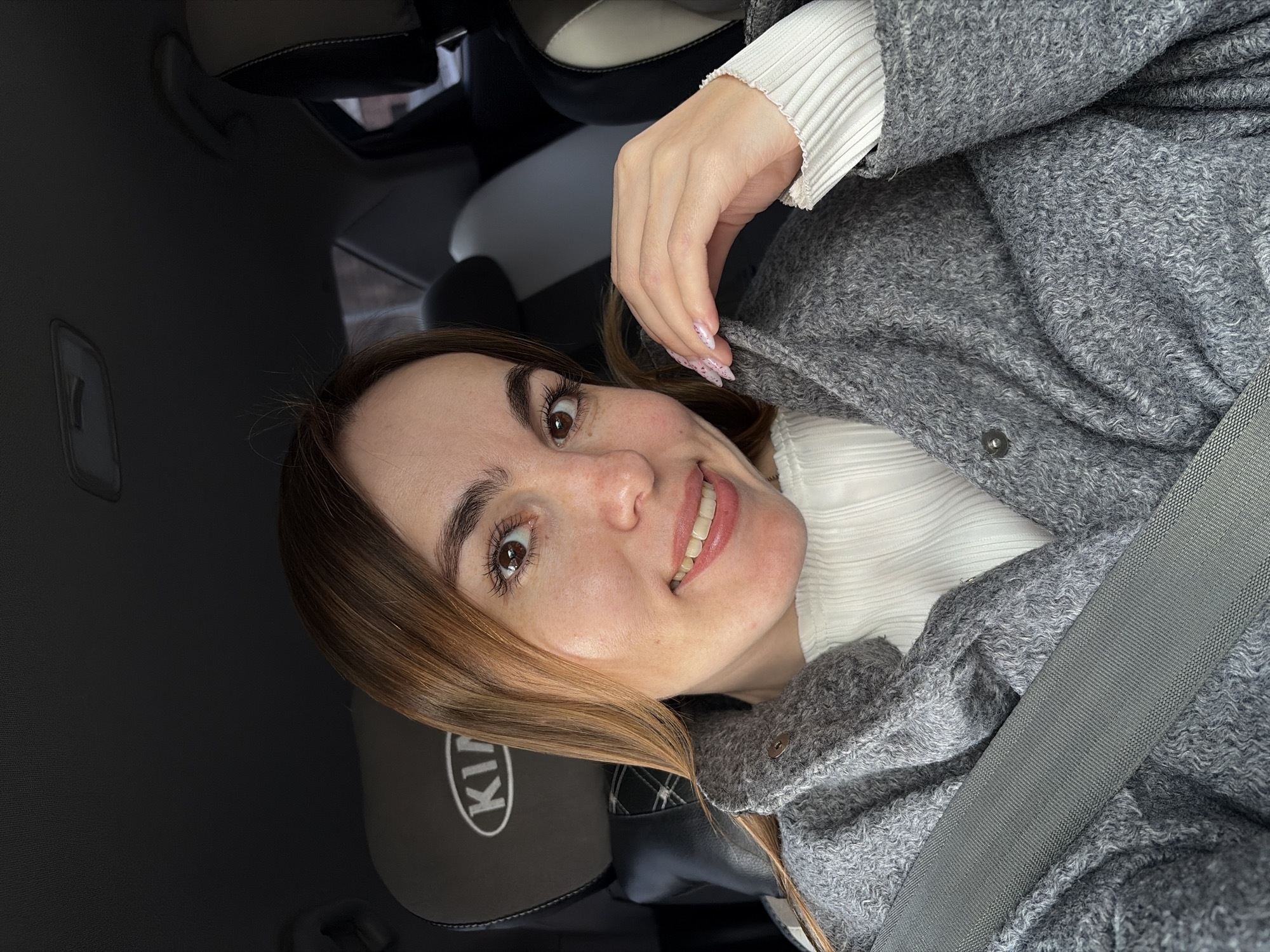 A woman is in the driver's seat of a Kia car, wearing a gray coat and white sweater, and smiling. The car's headrest is visible and has the Kia logo on it. She's wearing a seatbelt. The photo appears to be taken from within the car. There is no information to confirm the city of Izhevsk, Russia.