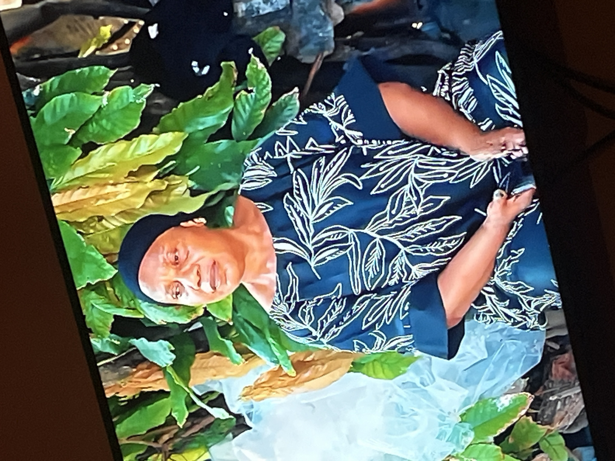 The image shows a medium shot of an adult Black woman seated outdoors, her body angled slightly to the left and her head turned to face the camera. She is wearing a dark blue headwrap and a dark blue, short-sleeved dress with a white leaf-like pattern. Her right hand is resting on her lap, and her left hand is partially visible near her waist. Behind her, lush green foliage with some yellowed leaves dominates the background. To the left of the woman, there appears to be a large, translucent white object, possibly plastic sheeting, and a portion of a wooden structure. To the right, a dark, possibly fabric, object and what looks like stacked stones are visible. The scene appears to be in a natural or semi-natural outdoor environment, likely within the city of Gura Top, Nigeria.