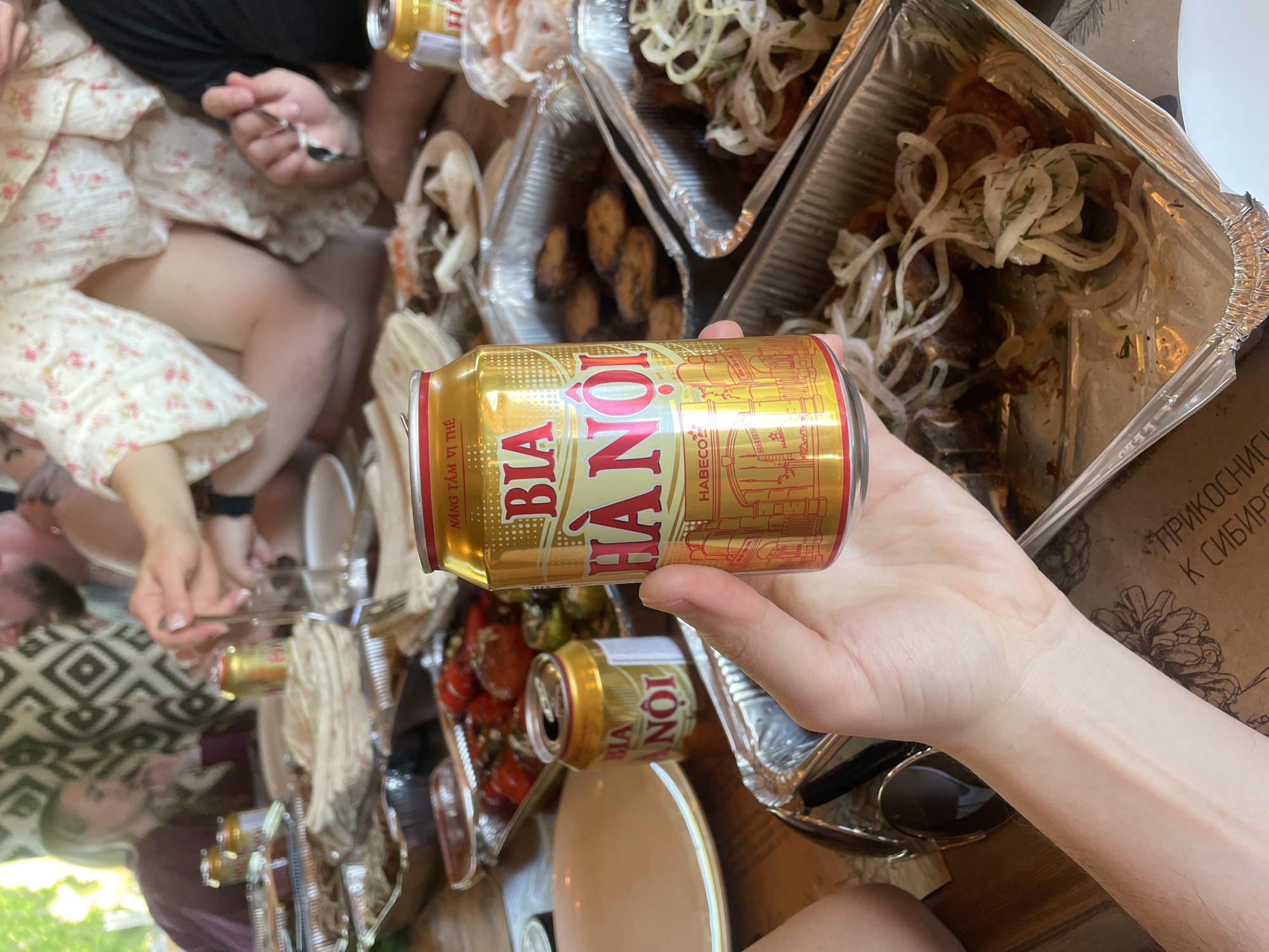 The image captures a casual gathering, likely a meal, with several people present, though most are only partially visible. The central focus is a hand holding a golden can of "BIA HÀ NỘI" beer, prominently displaying the brand name and "HABECO" logo. Another identical beer can lies on a brown table in the background.
The table is laden with food served in aluminum foil containers. One container holds what appears to be grilled meat or skewers generously topped with thinly sliced white onions and green herbs. Another container further back contains similar food, and a third container shows red, roasted vegetables, possibly tomatoes. To the left, a person wearing a floral-patterned dress or top is visible, with their legs and hands holding cutlery as if in the process of eating. Another person's arm and a patterned shirt can be seen further left. A pair of black sunglasses rests on the table.
In the lower right corner, a piece of brown paper or cardboard is visible with partially legible Russian text: "ПРИКОСНИС К СИБИРС", which translates to "Touch Siberia," providing a location cue consistent with the provided context of Дрокино, Russia. The scene suggests a relaxed daytime meal or picnic, with people enjoying food and beer.