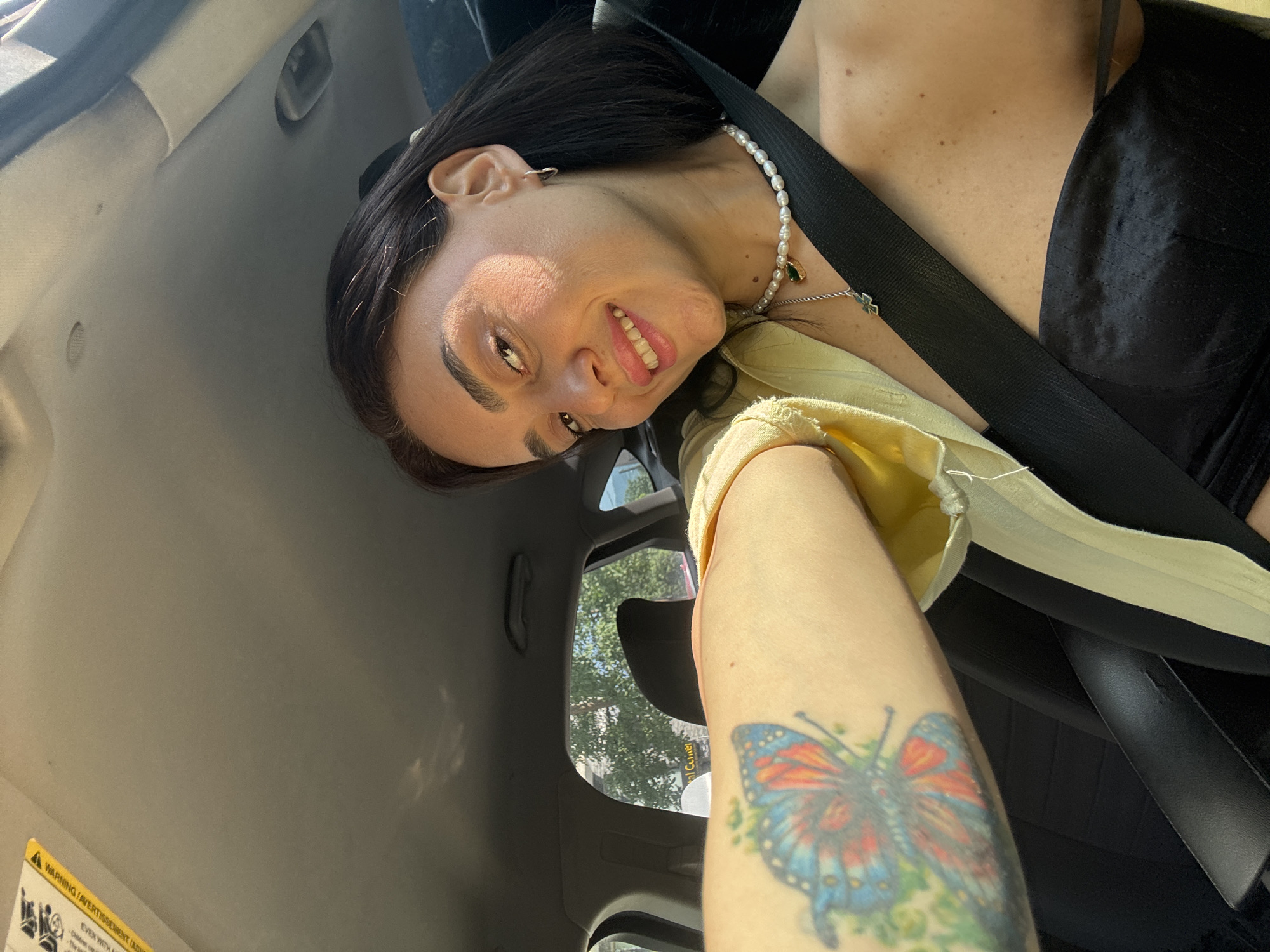 In an interior shot, a woman with dark hair and a tattoo of a butterfly on her arm is seen inside a vehicle. She is smiling at the camera and wearing a yellow shirt over a black top, a pearl necklace, and a seatbelt. The sun is shining, casting shadows on her face and the car's interior. Outside the car, trees and buildings are visible through the windows, suggesting an urban environment. The specific location of Tbilisi, Georgia, is not discernible from the visual information in the image.
