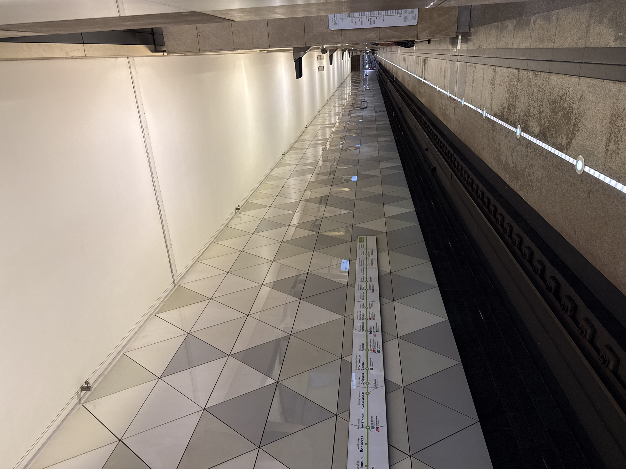The image shows a subway station platform in Moscow, Russia. The station features a modern design with a geometrically patterned wall made of white and grey triangular tiles. A long strip of illuminated signs is mounted on the wall, displaying a metro map and station names in Russian. The platform is empty except for a single person visible on the right side of the image, dressed in dark clothing, standing near a vertical information board. The tracks are visible in the foreground, with a train partially visible in the distance. The ceiling is illuminated with recessed lighting, creating a bright and clean atmosphere. The floor of the platform has a white line with circular illuminated markers, indicating the safe waiting area.