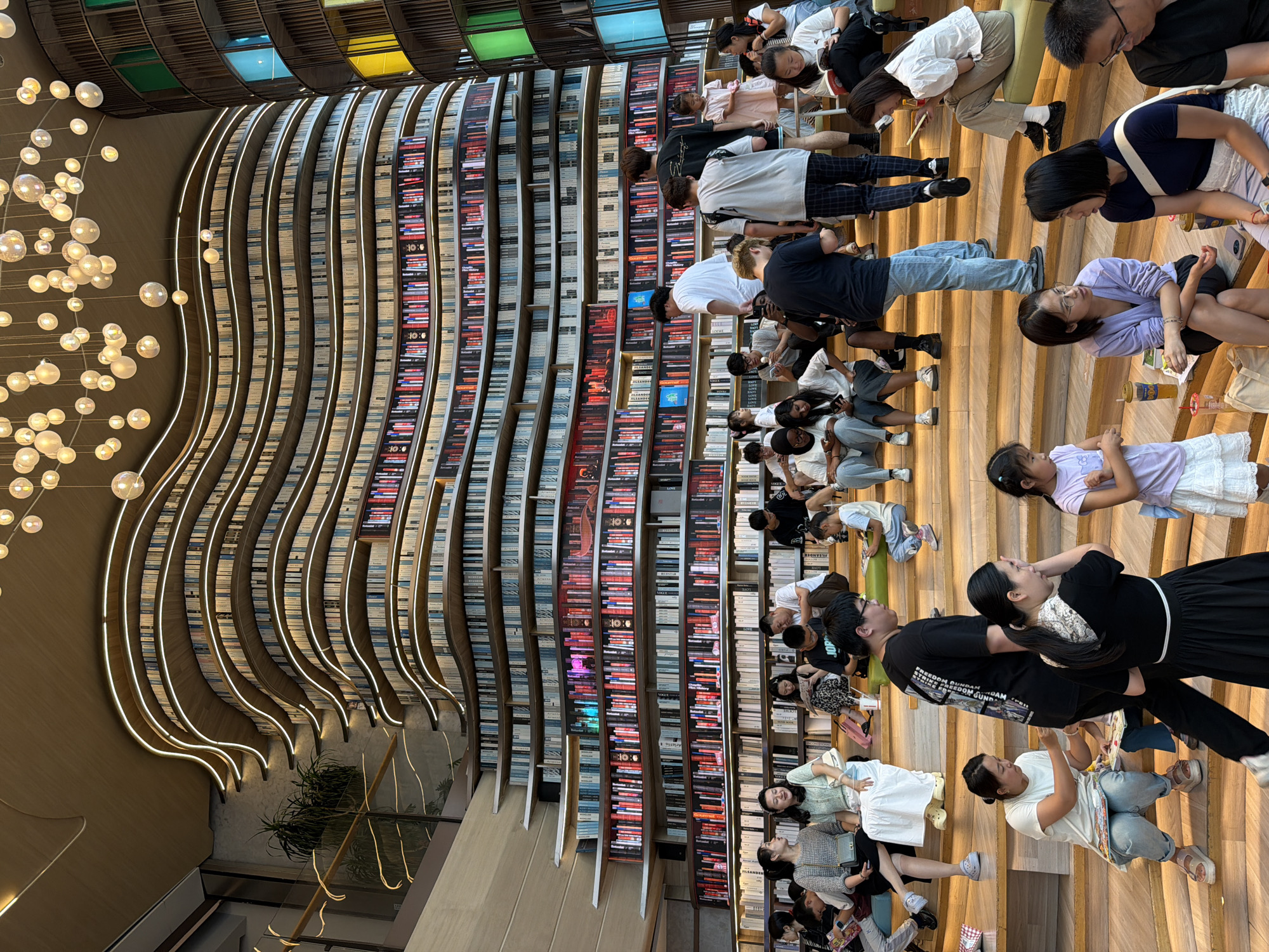 In the city of Liangjiang, China, a modern library features a striking, multi-tiered bookshelf design that curves and undulates like a wave. The bookshelves are illuminated with soft, strip lighting that accentuates their form. Numerous books are displayed on the shelves, with some sections featuring colorful spines or subtle colored lighting. A diverse group of people, including adults and children, are seated on wooden steps that lead up to the bookshelves. They are engaged in various activities such as reading, talking, and using their mobile phones. The overall atmosphere appears relaxed and communal, suggesting a popular public space for leisure and learning.