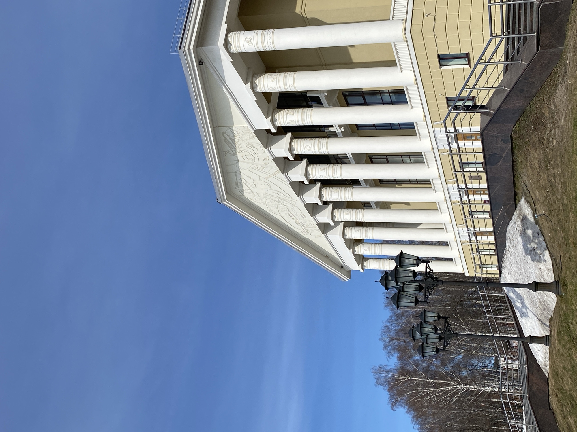 The image shows a section of a building with classical architectural elements against a bright blue sky. The building has a yellow facade with white columns. The columns appear to support a triangular pediment adorned with bas-relief carvings. Windows are visible behind the columns. A black marble staircase with a metal railing runs alongside the building. The ground near the building includes patches of snow and brown vegetation. Vintage-style lampposts stand near the trees. The image is taken in the city of Votkinsk, Russia.