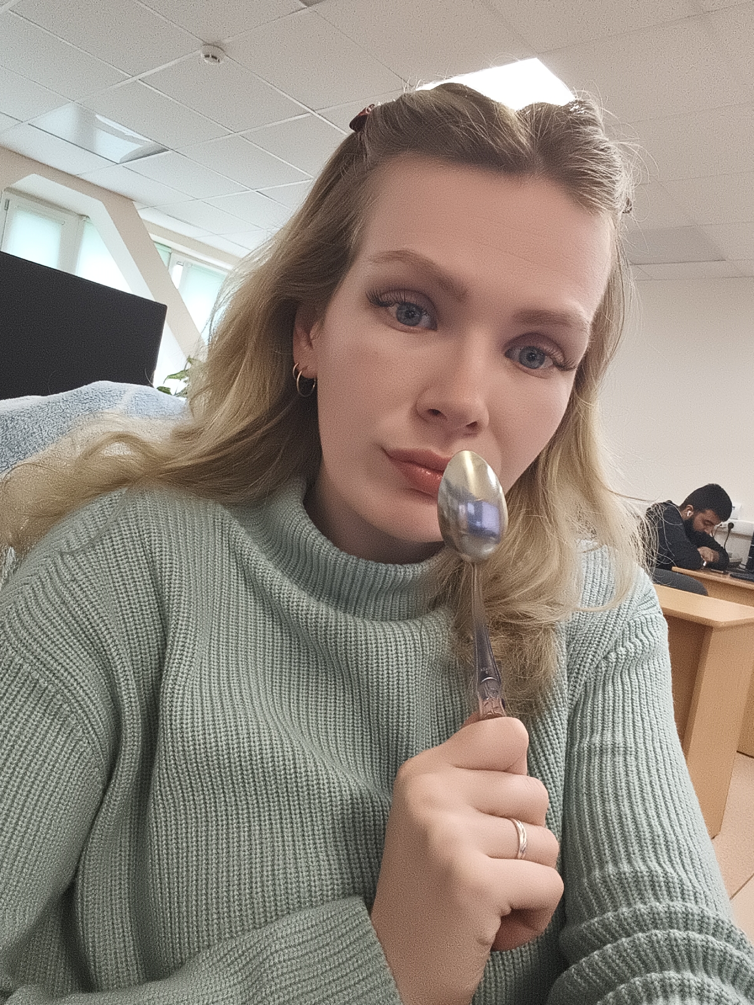 A young, fair-skinned woman with long blonde hair is holding a silver spoon up to her face. She is wearing a light green, ribbed sweater, and a simple ring on her ring finger. Her eyes are blue, and she has long eyelashes.
In the background, a man with dark hair is seated at a desk, facing away from the camera. The room appears to be an office or classroom setting, with desks, chairs, and a window. The windows reveal a cityscape that is consistent with the architecture and urban design of Izhevsk, Russia.