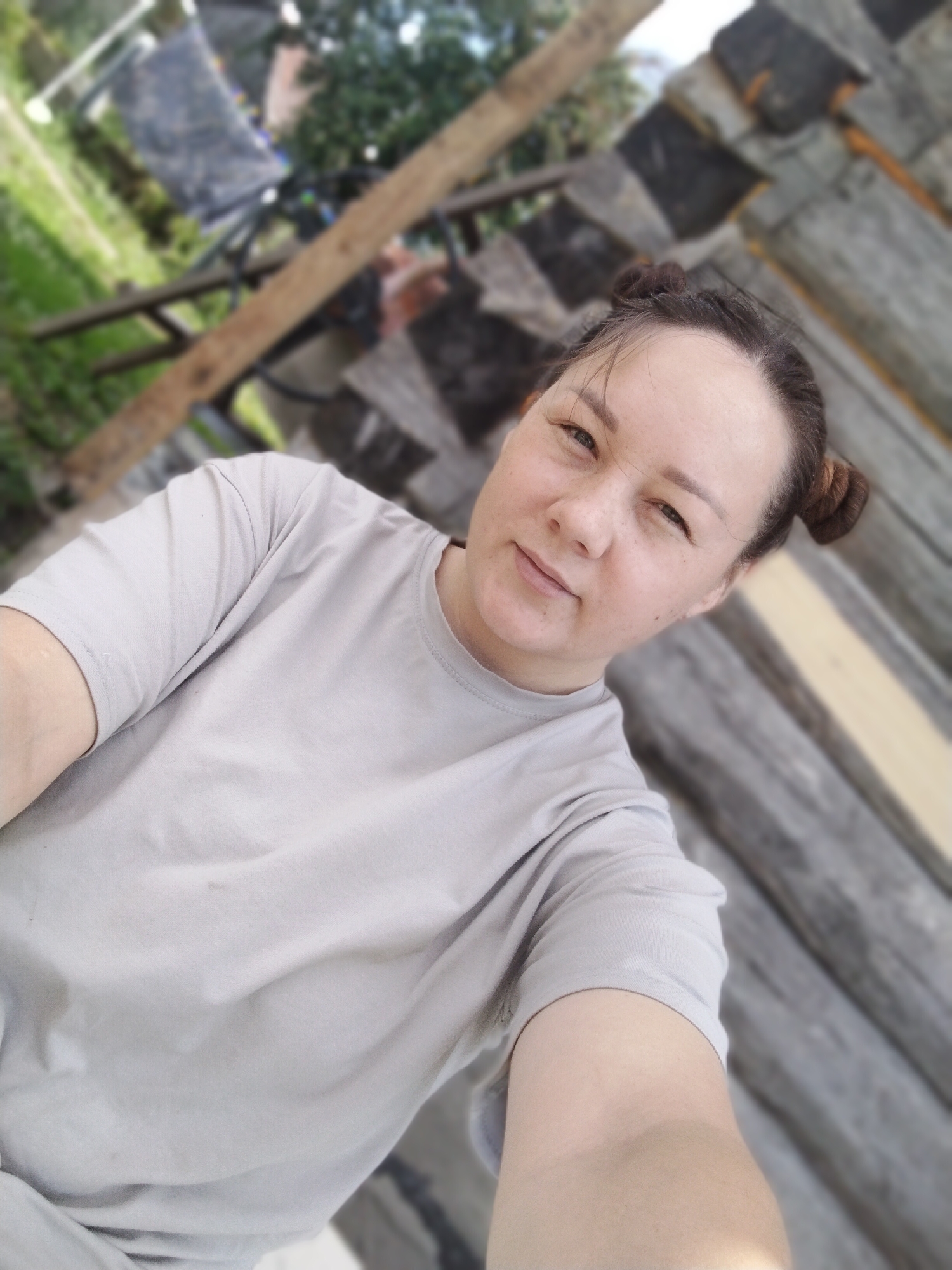 The image shows a woman with her hair styled in two buns, wearing a light grey t-shirt. She is outdoors, with a wooden structure to her right and greenery and what appears to be some construction materials in the background. The environment suggests a rural or semi-rural setting. The lighting indicates it is daytime. The image is a selfie, taken from the woman's perspective.