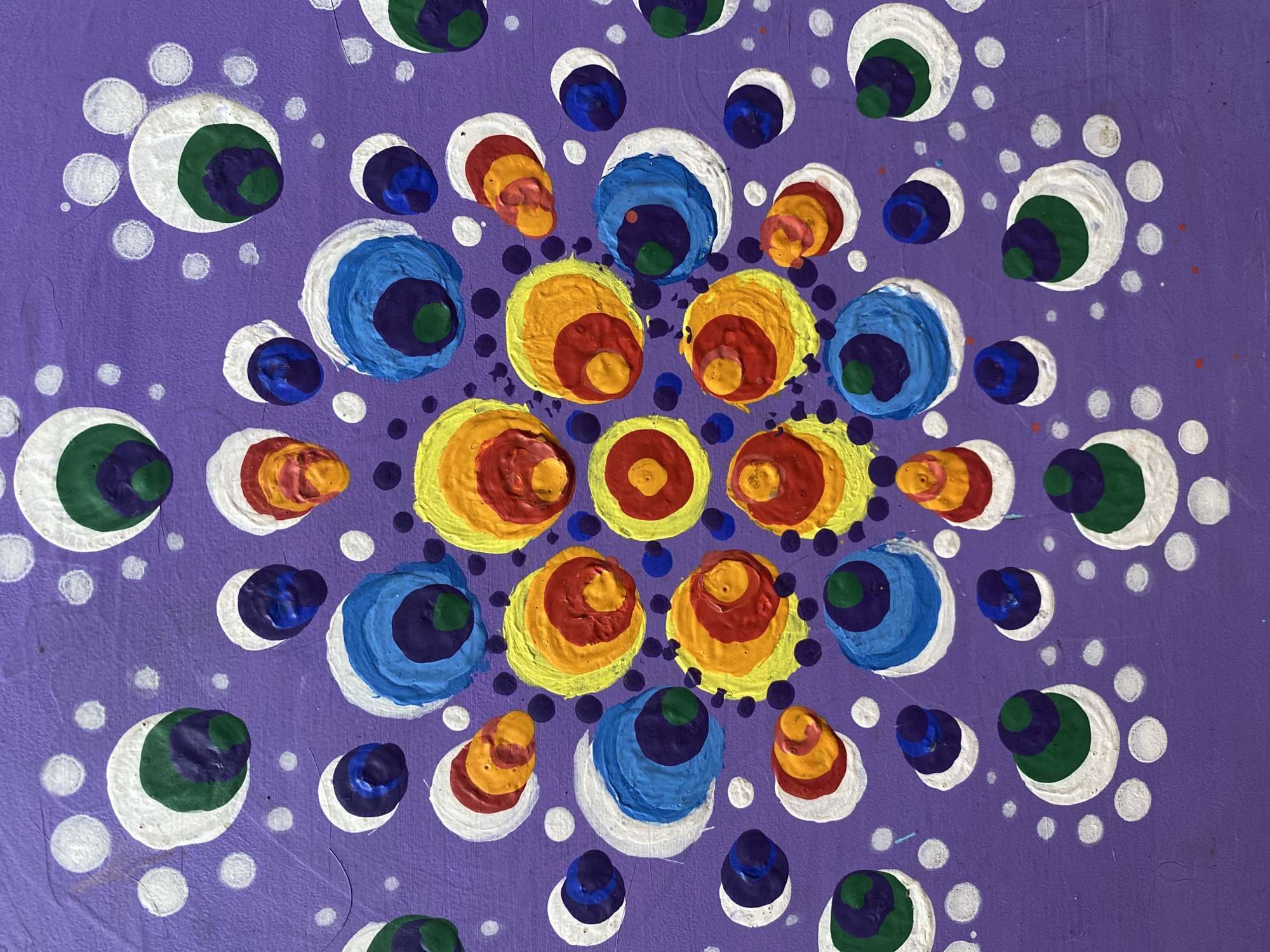 This is a close-up, top-down photograph of a piece of artwork on a textured purple surface, possibly canvas or wood. The artwork is a mandala-style design created with acrylic paints. The design consists of concentric circles of various colors arranged in a radial pattern. Dominant colors include white, green, dark blue, light blue, yellow, red, and orange. Small white dots are scattered around the larger circular elements, filling the purple background. The paint appears to be applied thickly, giving the circles a raised, textured appearance. There is no indication of the artwork's location in Zelenodol'sk, Russia, as this is a photograph of a standalone artwork.