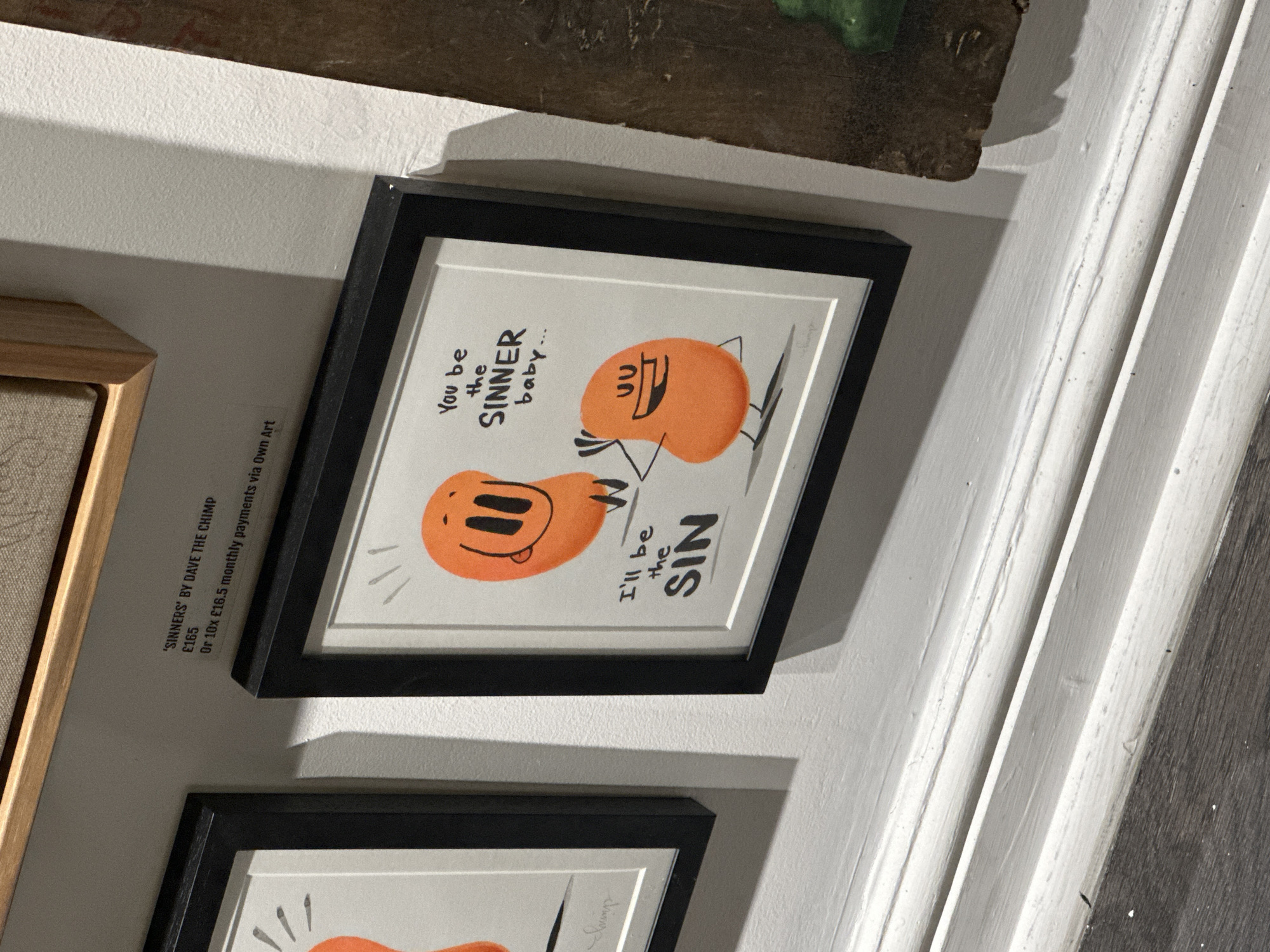 This is a close-up, rotated photograph capturing framed artwork displayed on a white wall, likely within an art gallery or shop in London, United Kingdom. The prominent piece is a framed print featuring two orange, blob-like cartoon characters against a light background. One character, with large eyes and a smile, has a speech bubble that reads, "You be the SINNER baby...". The other character, depicted with a mischievous expression and a tongue sticking out, has text next to it saying, "I'll be the SIN". The artist's signature, "Swimp" (likely "Chimp"), is visible on the artwork.
To the left of the main artwork, a small label provides details: "'SINNERS' BY DAVE THE CHIMP". The price is listed as "£165", with an alternative payment option of "Or 10x £16.5 monthly payments via Own Art". Part of a second, similar framed artwork is visible below the main piece. The wall itself is white with a subtle texture, and a decorative white architectural molding runs down the right side of the image. The setting appears to be an indoor commercial space for art, highlighting the playful and slightly suggestive theme of the cartoon artwork.