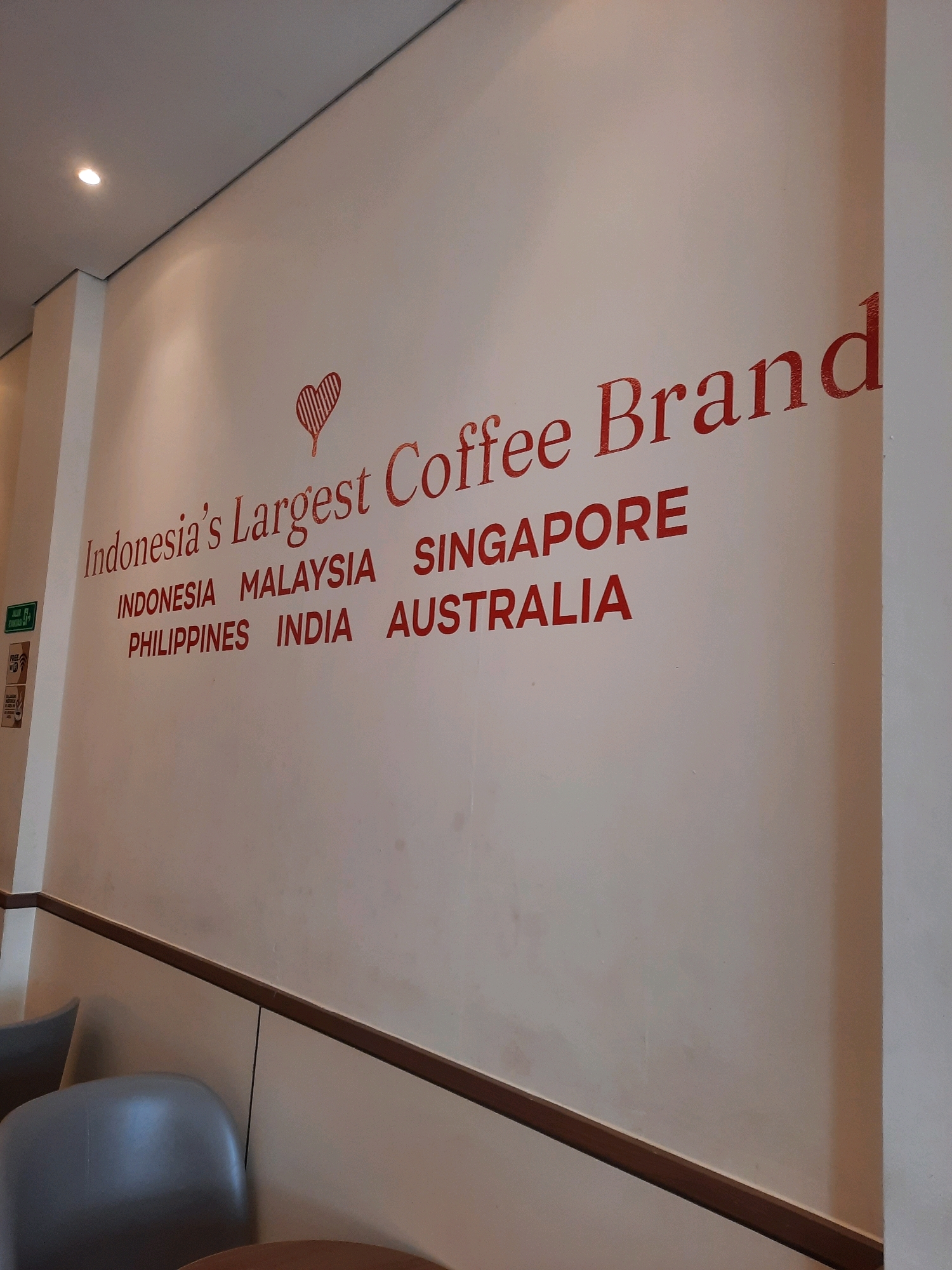 The image shows a red text written on a white wall inside a cafe. The text reads "Indonesia's Largest Coffee Brand" and below it, a list of countries: "INDONESIA MALAYSIA SINGAPORE PHILIPPINES INDIA AUSTRALIA". There are also some signs on the left wall of the cafe, including a green sign with white text "Jalan" and a small sign that says "FREE WIFI". The cafe appears to be located in East Jakarta, Indonesia, as indicated by the text on the wall and the presence of the word "Jalan" which means "street" in Indonesian. The overall scene depicts a branded interior of a coffee shop.