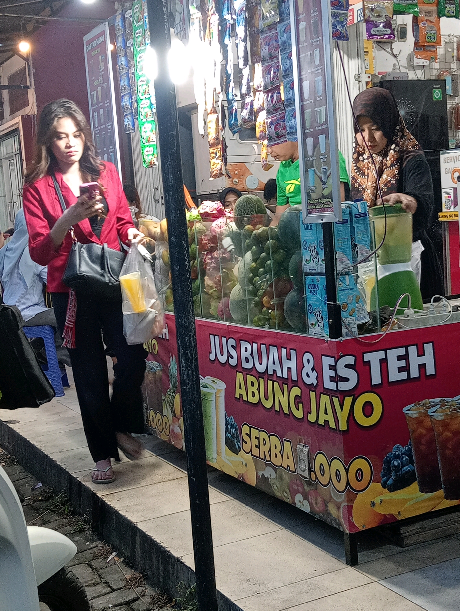 In Bandar Lampung, Indonesia, a street food stall named "JU BUAH & ES TEH ABUNG JAYO" is operating at street level. The stall features a bright red canopy with large white lettering advertising "JU BUAH & ES TEH ABUNG JAYO" and "SERBA 1.000" along the side, which translates to "Fruit Juice & Ice Tea Abung Jayo" and "Everything 1,000". The stall displays an assortment of fresh fruits, including melons, oranges, and grapes, and also sells bottled drinks, as evidenced by the presence of milk cartons. A blender is visible on the counter, suggesting fresh juice preparation. Two women are present: one in the foreground wearing a red jacket and black pants, looking at her phone and holding a plastic bag with a beverage, and another woman behind the counter, wearing a headscarf and a patterned brown and beige shawl, operating the stall. In the background, a menu board with Indonesian text is displayed, and various packaged goods hang from the stall's structure. The environment appears to be a busy outdoor market or street setting, with a light post illuminated near the stall. The ground is paved with concrete tiles.