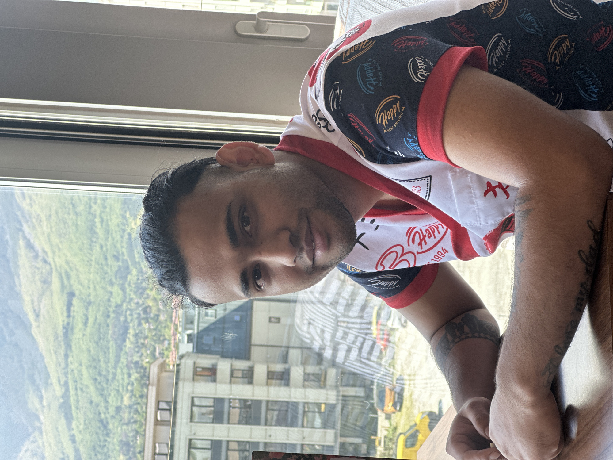 The image shows a man sitting at a table next to a window. The window reflects the city of Sofia, Bulgaria, including a view of green trees and buildings. The man is wearing a t-shirt that is white with a red collar and sleeves with a repeating print of the word "Happy". He has dark hair, brown eyes and facial hair. He has multiple tattoos on his arms, including a wristband on his left arm and a tattoo on his right arm that reads "love my dad".