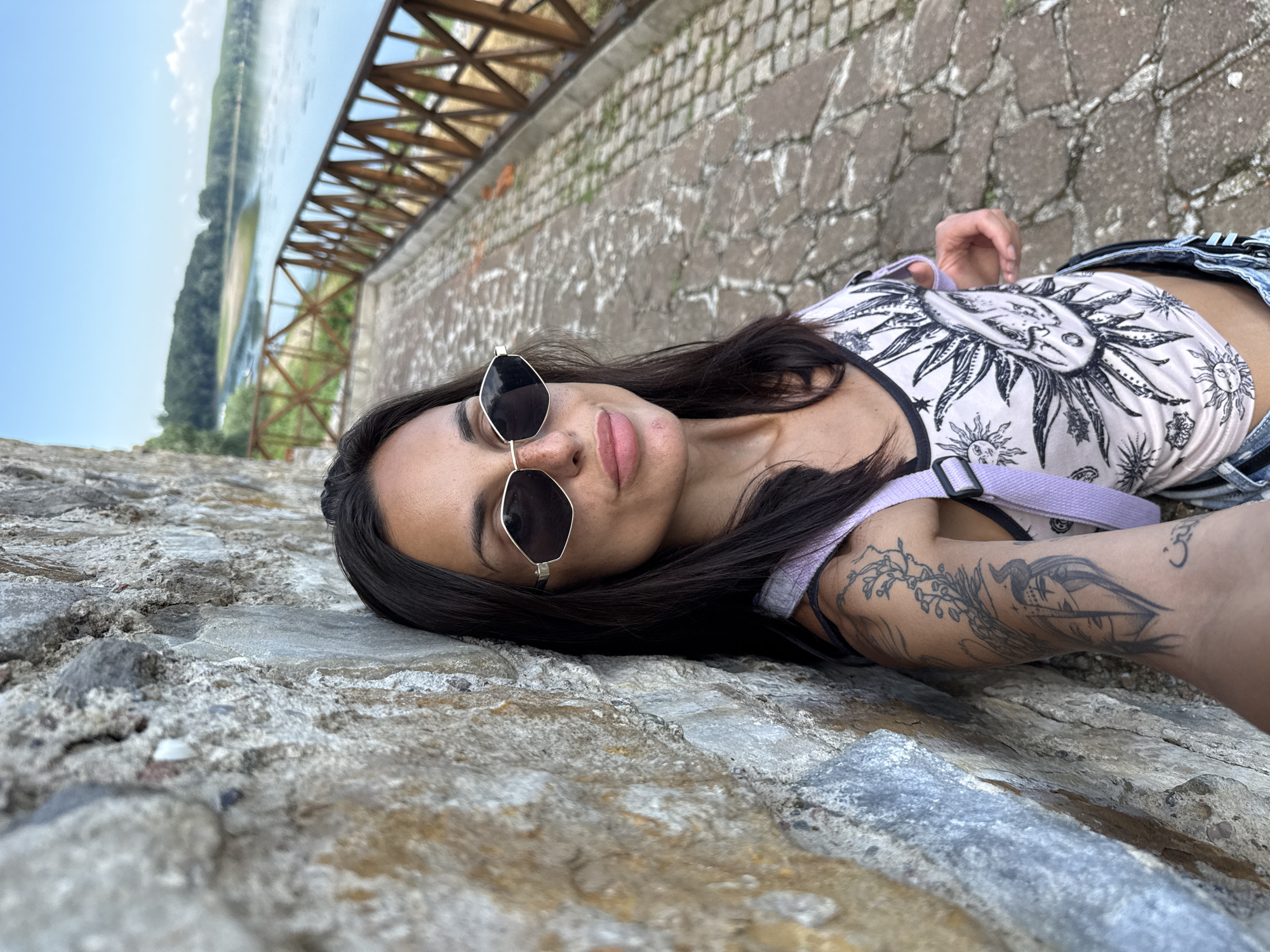 A woman with dark hair and sunglasses leans against a stone wall. She is wearing a tank top with a celestial print and denim shorts. A light purple strap is visible over her shoulder, part of a bag. Her left arm, which is visible, has tattoos. She is in an outdoor setting with a paved pathway and a wooden railing. In the background, there is a body of water and a treeline under a blue sky with some clouds. This location is identified as Belene, Bulgaria.