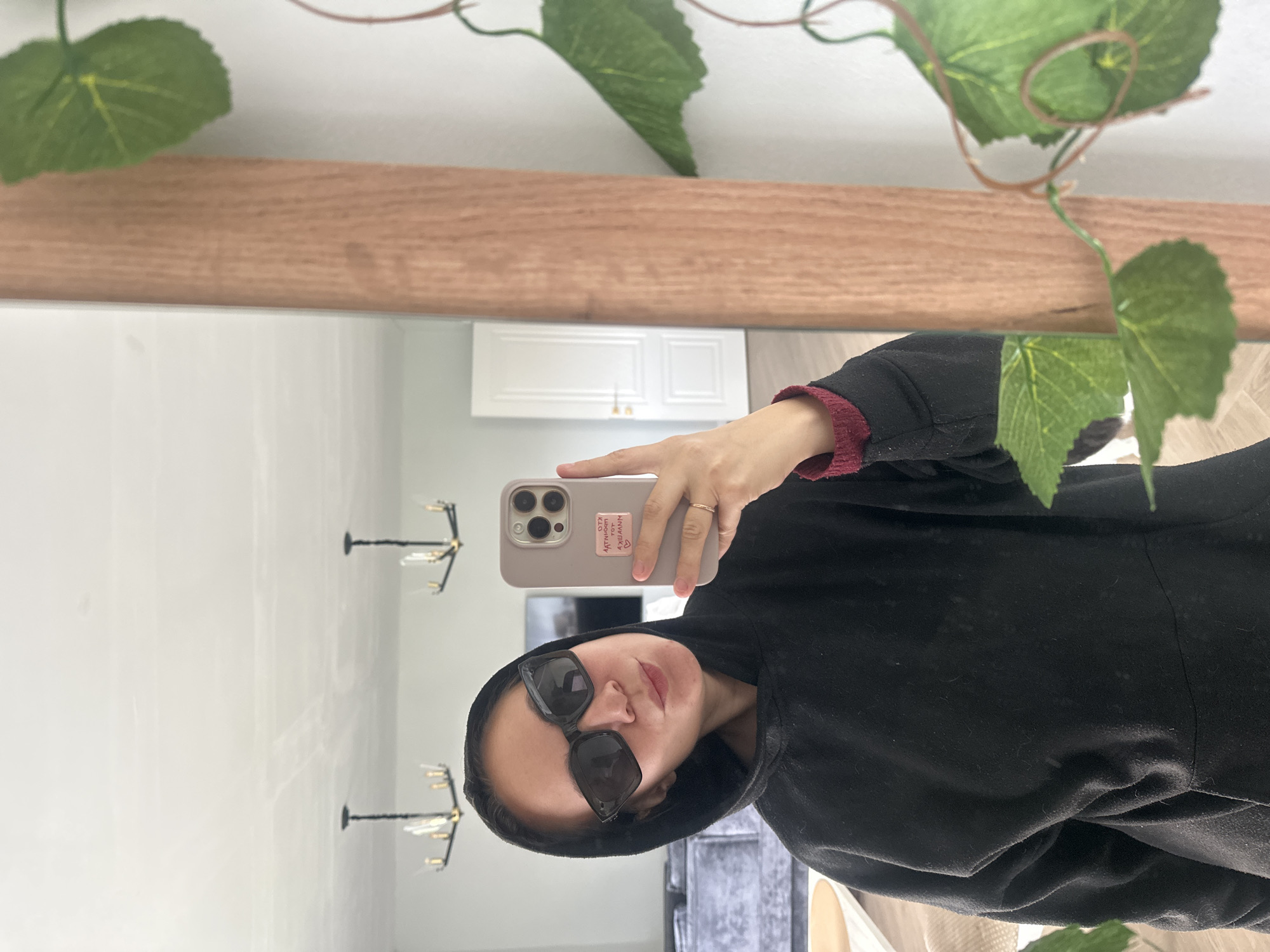 An individual is captured in a mirror selfie within a modern residential interior in Kharnavy, Russia. The subject, an adult, wears a black hooded sweatshirt with the hood up and black rectangular sunglasses. They are holding a light grey smartphone with a multi-lens camera array in their right hand, positioned to capture the reflection. A gold-toned ring is visible on the subject's right ring finger. The phone case features a white sticker with mirrored dark text. The subject's lips are slightly puckered.
The mirror has a light brown wooden frame and reflects a brightly lit room. In the background reflection, two modern black chandeliers with multiple light sources are suspended from a white ceiling. Further details visible in the reflection include a grey sectional sofa, part of a light-colored round table, and a white interior door with a gold handle. To the right of the mirror, an artificial green vine with several leaves and brown stems is draped vertically against a white wall. The room's walls and ceiling are white, with subtle seams visible on the ceiling.