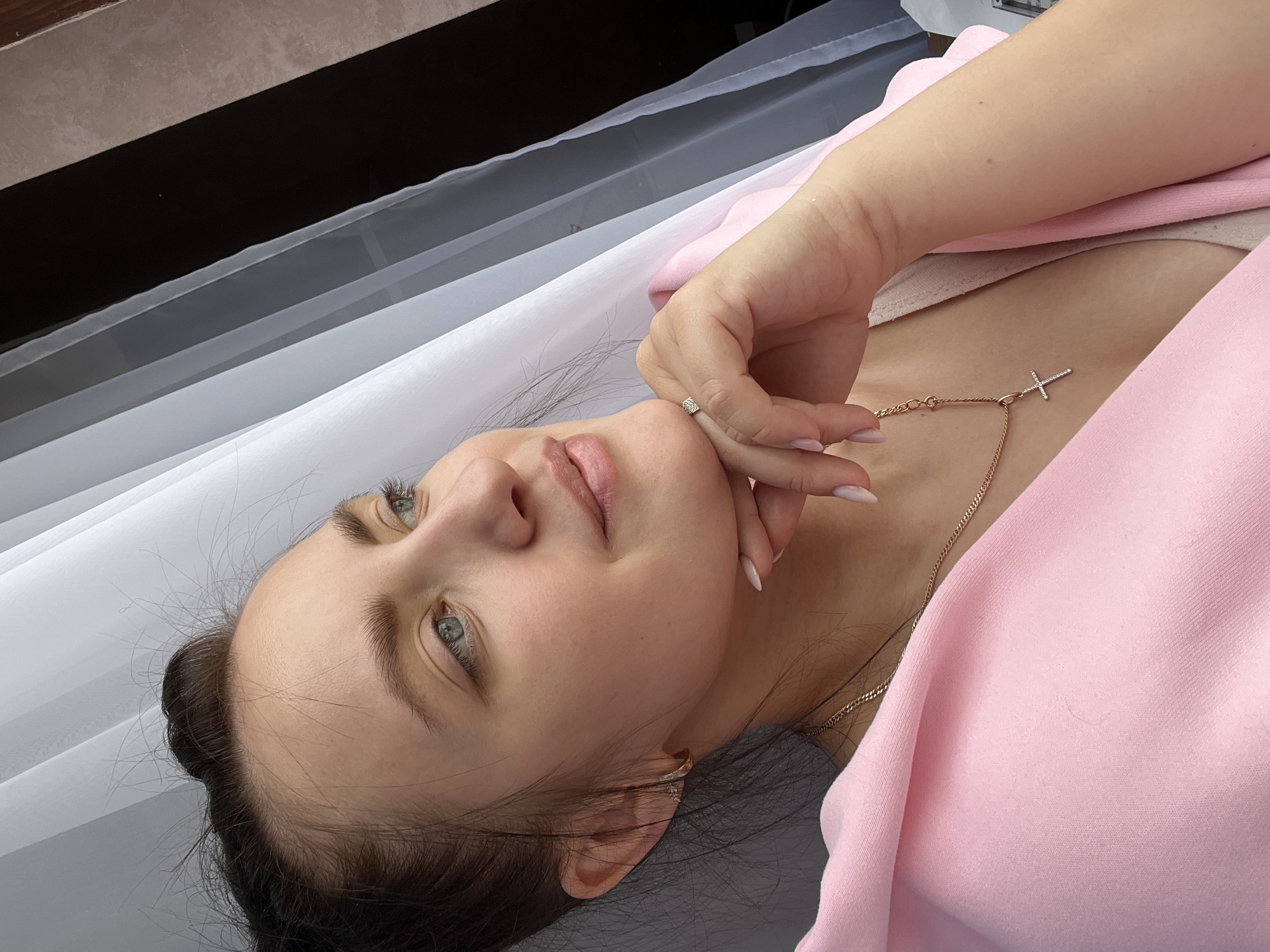 A woman with fair skin, green eyes, and dark hair lies on her back, wearing a pink top and a gold necklace with a cross pendant. Her right hand is raised, with fingers touching her cheek. She wears a ring on her ring finger. The background includes a white cloth and a dark gray surface. The location is Марковский, Russia.