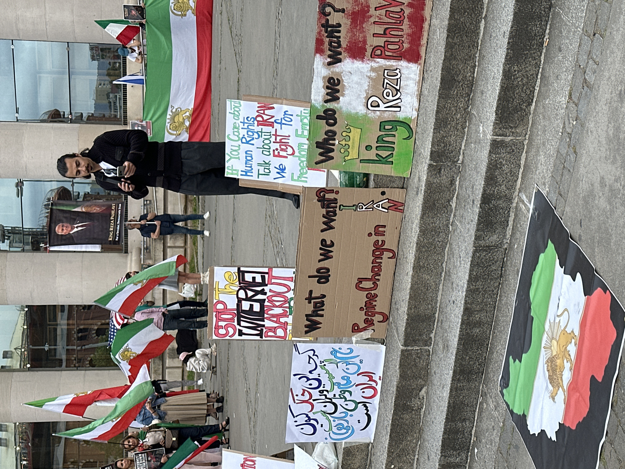 A group of protesters are gathered on the steps of a building, presumably in Barcelona, Spain, based on the Catalan flag visible in the background. They are holding signs and Iranian flags. The signs bear messages such as "If you care about human rights, talk about Iran we fight for freedom,” “King Reza Pahlavi,” and “Stop the internet blackout.” There is one man wearing a dark sweater in the middle of the scene, looking at his phone.