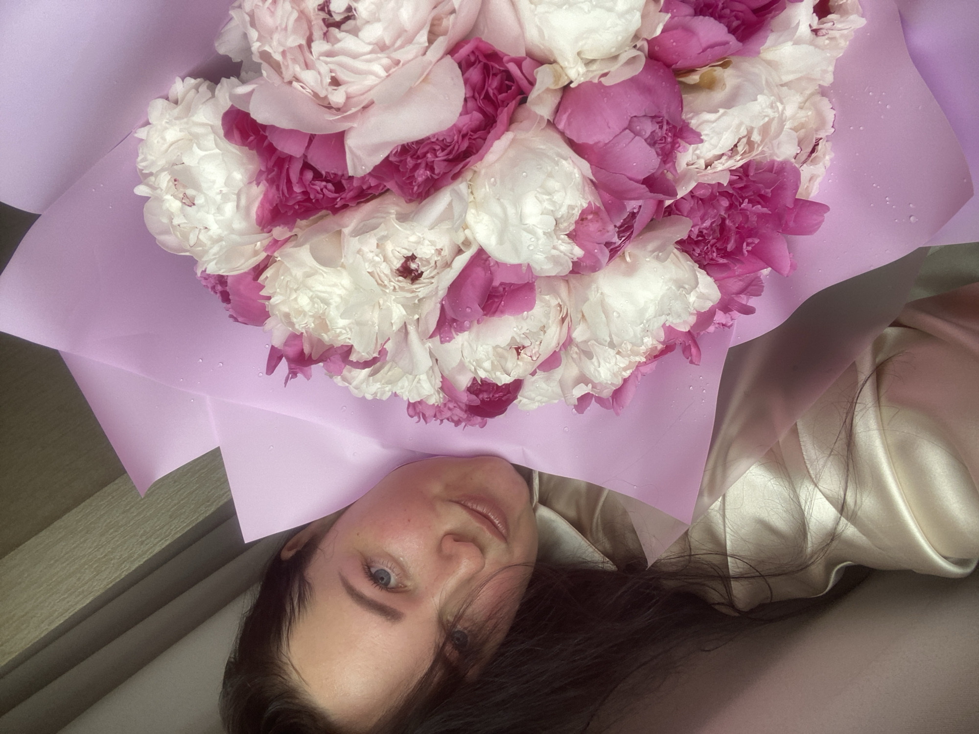 A woman with light skin, blue eyes, and dark hair is holding a bouquet of white and pink peonies wrapped in light purple paper. The woman is wearing a light-colored silky garment. The location is not discernible from the image.