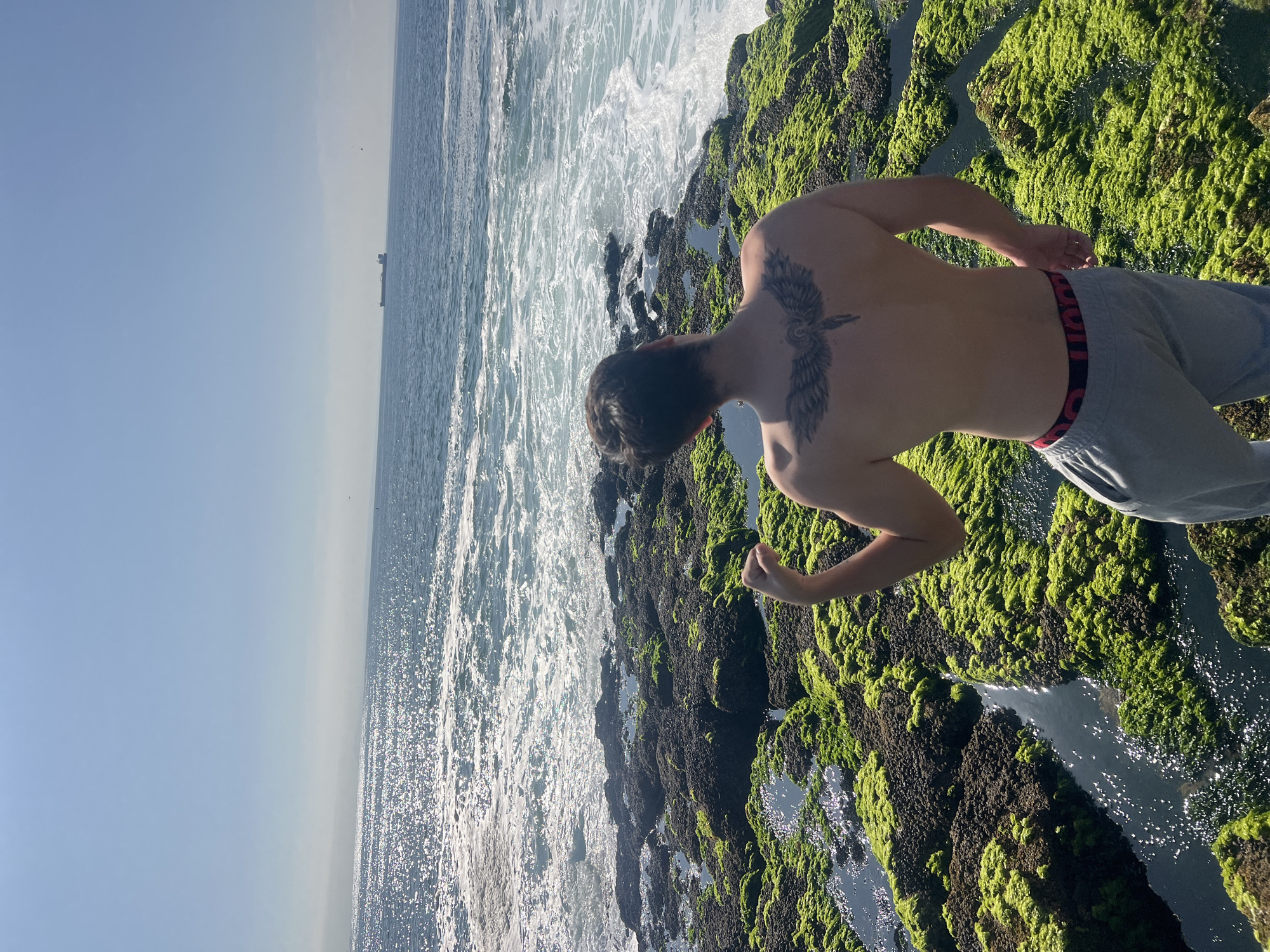A photograph taken from a high angle depicts a person standing on a rocky, moss-covered shore next to the ocean. The person, a male based on physique and apparent clothing, is shirtless, wearing grey pants with a visible red waistband. He has a tattoo of a bird or winged creature on his upper back. The ocean water is choppy with whitecaps, reflecting sunlight. A ship is visible on the horizon. The rocky area is densely covered in green moss and algae. This location is likely on the coast in Arica, Chile.
