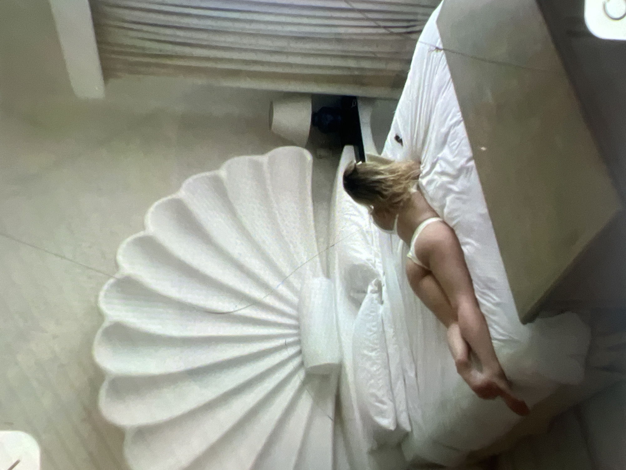 A woman in white lingerie lies on a bed in a room. The bed has a large, white, shell-shaped headboard. There are two pillows on the bed, and the woman is positioned on her stomach. She has blonde hair and is facing away from the camera. A lamp with a white shade sits on a nightstand next to the bed, and a curtain hangs in the background. The floor appears to be tiled. There is no indication of the location being in Kara-Dıykan, Kyrgyzstan.