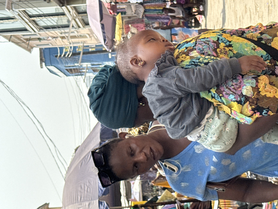 The scene depicts a bustling street in a market area of Lagos, Nigeria, under a bright, possibly hazy sky. In the foreground, two dark-skinned adult women and an infant are present. The woman on the left, an adult female, wears a light blue patterned v-neck top, has dark sunglasses perched on her head, a small nose ring on her right nostril, and a white wired earbud in her right ear connected to a small white clip-on device. Her mouth is slightly open, suggesting speech or a reaction, and her gaze is directed towards the second woman.
The second adult female, positioned centrally and partially obscured, wears a dark teal headwrap and small gold stud earrings. She is carrying an infant on her left side/back, secured by a brightly colored, multi-patterned fabric with floral designs in yellow, pink, blue, and green. The infant, wearing a grey long-sleeved top with ruffled trim and light green patterned bottoms, appears to be asleep, resting its head on the carrier's shoulder with eyes closed. Small gold stud earrings are visible on the infant.
The background reveals a vibrant market environment. On the left, a blue and white striped umbrella shades a stall. On the right, another light-colored umbrella covers a stall displaying colorful fabrics. Behind the market stalls, multiple-story buildings are visible, including a prominent blue-painted structure and a lighter-toned building with numerous windows. Overhead, numerous black electrical or utility wires crisscross the sky. The ground appears to be a light, dusty surface. Other indistinct figures are present further in the background, including a person in a white tank top seated on the far right.