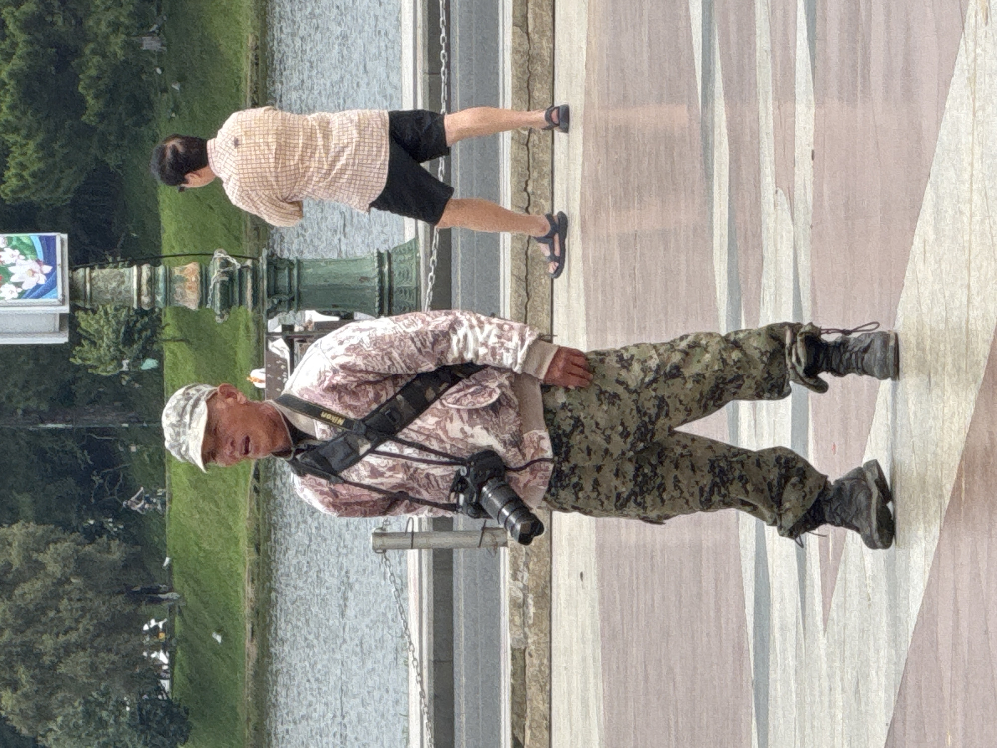 The image captures a street scene in Da Lat, Vietnam, near Xuan Huong Lake. In the foreground, a man with a camouflaged cap and pants stands facing forward, wearing a patterned jacket and dark boots. A camera with a lens is slung across his chest. Behind him, another man in a checkered shirt and black shorts is visible, with his back to the camera, looking away. The background shows a body of water, a grassy embankment, and trees, indicative of a park or lakeside setting. A decorative lamppost stands between the two men.