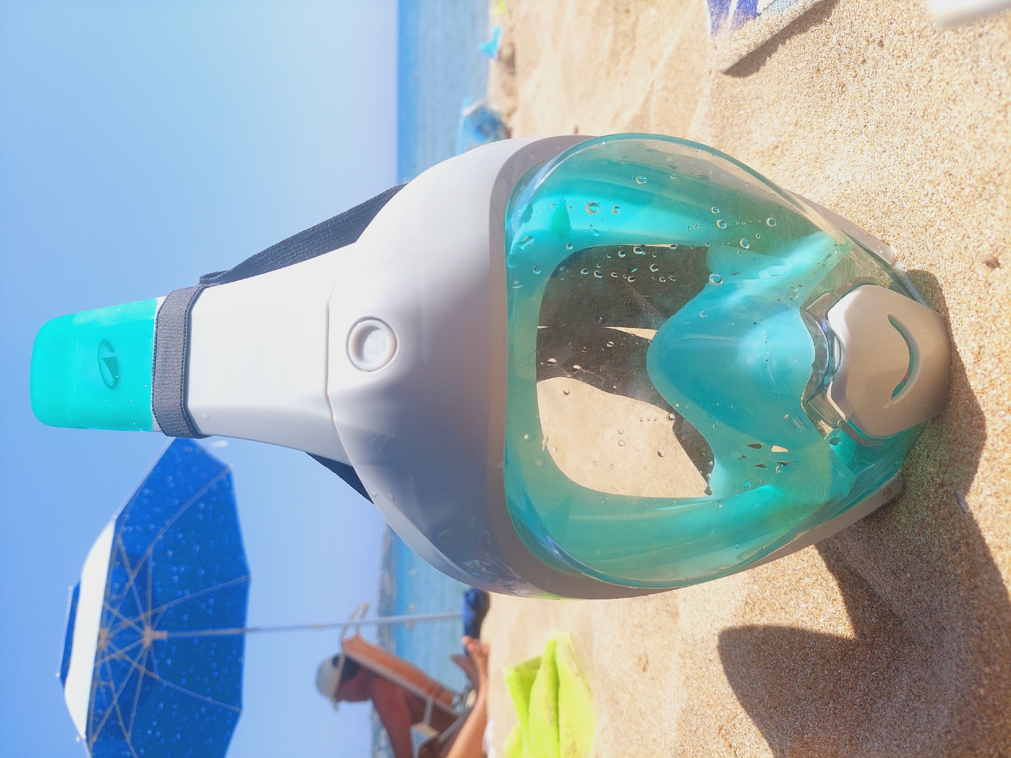 A full-face snorkel mask rests on the sandy beach in Sozopol, Bulgaria, with the turquoise sea and a blue and white striped beach umbrella in the background. The mask is white and gray with a turquoise lens. Water droplets are visible on the lens, suggesting it has been used or is ready for use. In the background, a person sits in a beach chair under the umbrella, facing away from the camera, and appears to be relaxing. The sky is clear and blue, and the sun is shining, creating a bright and summery atmosphere.