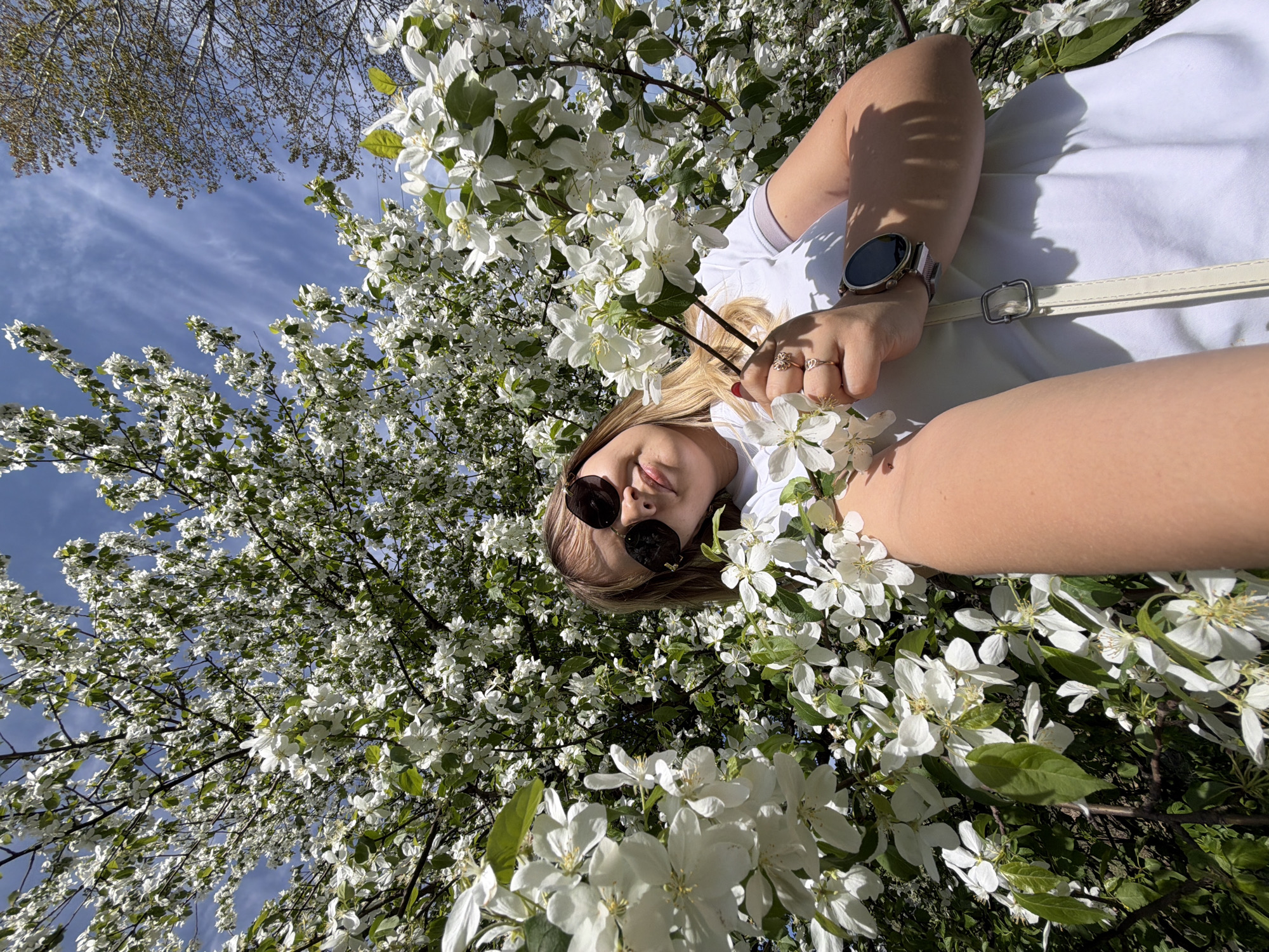 The image shows a fair-skinned woman wearing sunglasses and a white t-shirt lying amongst a tree with white blossoms under a blue sky. The woman is wearing a watch and rings, and has a white strap over her shoulder. The location is likely Chaikovsky, Russia.