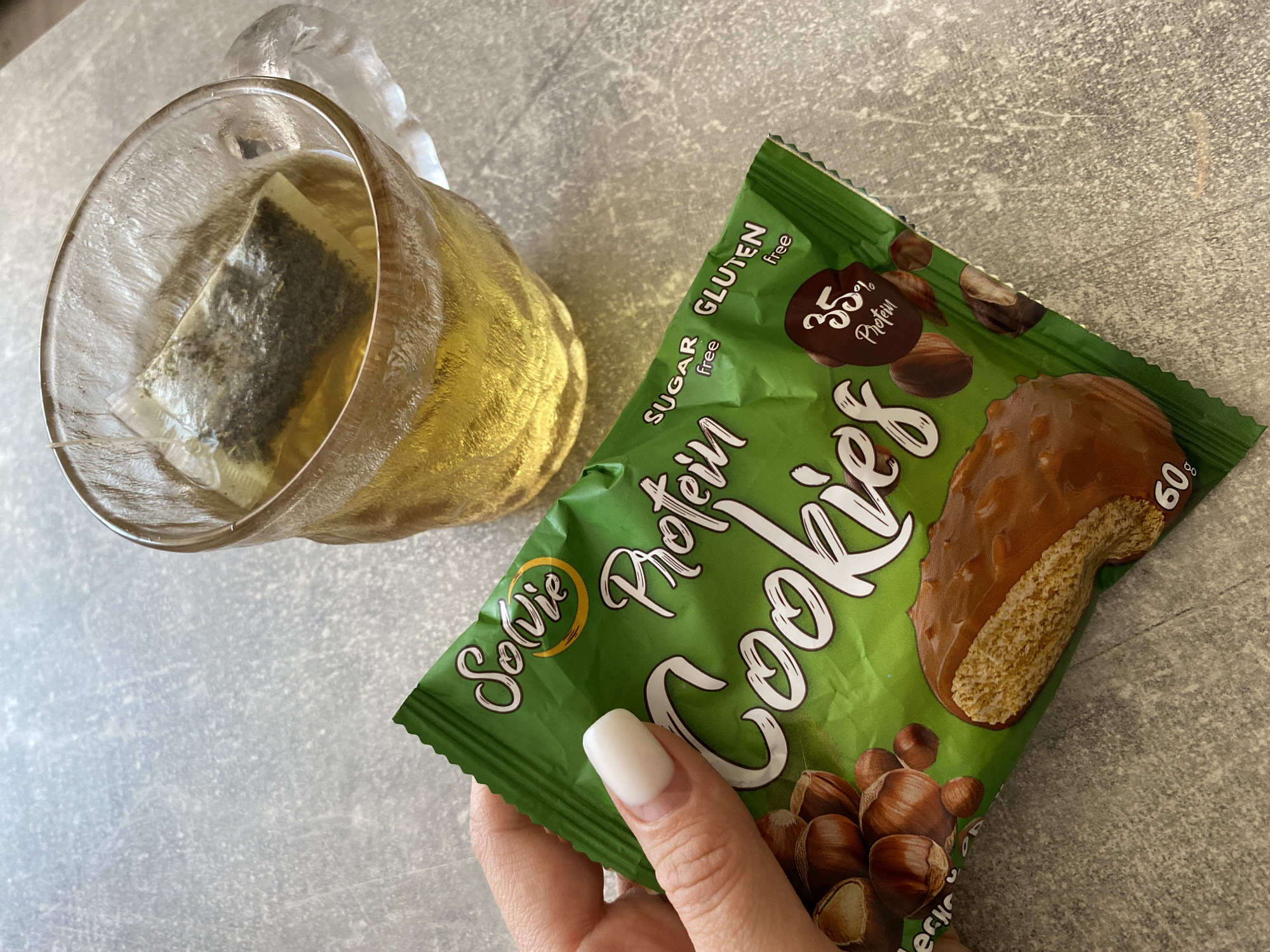 A photograph depicts a still life scene featuring a cup of tea and a package of "Solvie" Protein Cookies. The tea is in a clear glass mug with a tea bag steeping in the golden liquid. The cookie package is green and prominently displays the words "Solvie Protein Cookies" in white lettering. Additional text on the package indicates "sugar free" and "gluten free," with a claim of "35% protein." An image of a chocolate-coated cookie and hazelnuts is also visible on the package. A hand with a light-colored nail polish holds the cookie package. The objects rest on a surface with a mottled gray pattern. The image was taken in the city of Chaikovsky, Russia.