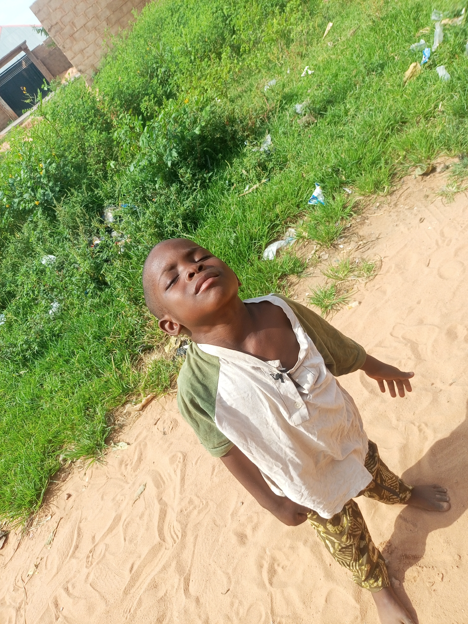 The image depicts a young boy with his eyes closed, facing upwards, standing in a sandy area next to a patch of overgrown grass. The boy is wearing a light-colored shirt with green sleeves and patterned green pants. In the background, there is a brick building under construction and more dense green vegetation, some of which appears to be overgrown. Some scattered debris, possibly plastic bags, is visible in the grass. The overall setting suggests an outdoor environment in a developing area, consistent with parts of Tempure, Nigeria. The lighting indicates it is daytime, with shadows suggesting the sun is positioned overhead.