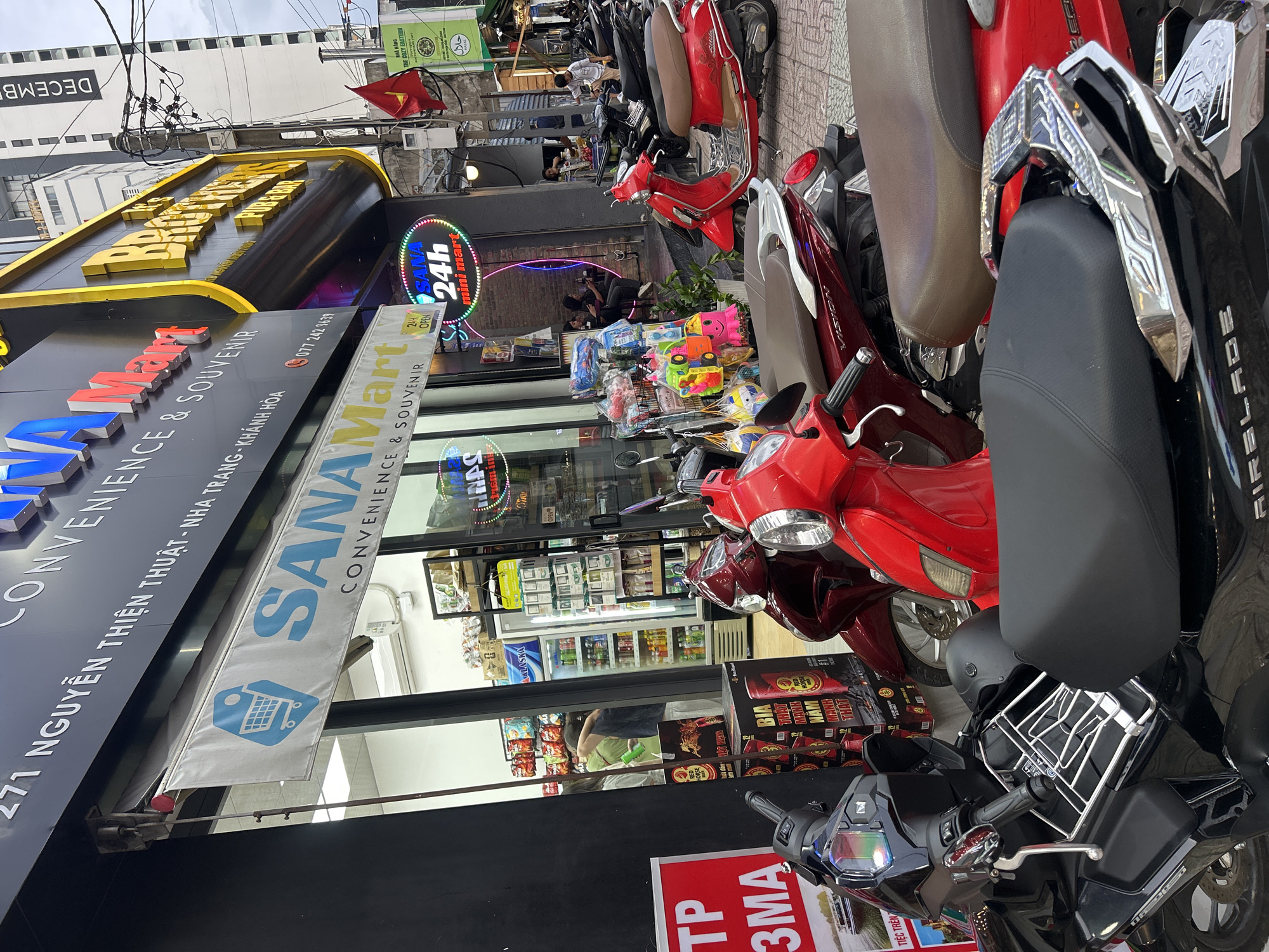 The image captures a vibrant street scene in Nha Trang, Vietnam, dominated by parked motorbikes and storefronts. In the foreground, numerous motorbikes, primarily red and black scooters including one with "AIRBLADE" text, are tightly parked along the sidewalk.
The central focus is the "SANA Mart" convenience store, identifiable by its prominent blue and white signage. A banner with a shopping cart icon hangs above the entrance, and neon signs advertise "24h OPEN" and "mini mart." The store's windows reveal refrigerators stocked with beverages, including "ALASKA" branded items, and shelves displaying various products. Outside the store, a colorful array of toys is displayed, and large red boxes of "RED HORSE" branded beer ("BIA HÒI ĐỨC MẠNH") are stacked near the entrance.
Further into the scene, several people are visible, some sitting at tables in an outdoor area, suggesting a cafe or restaurant. A sign for "NHÀ HÀNG THE COZY EASTERN" with a "حلال" (Halal) symbol is also present. The sky above appears overcast, and dense electrical wires crisscross overhead. Key location information is visible on signage, including the address "271 NGUYỄN THIỆN THUẬT - NHA TRANG - KHÁNH HÒA" and a phone number "077 242 1659." Another partial sign reads "DECEMB" on a nearby building. The scene depicts a typical busy commercial street during the daytime.