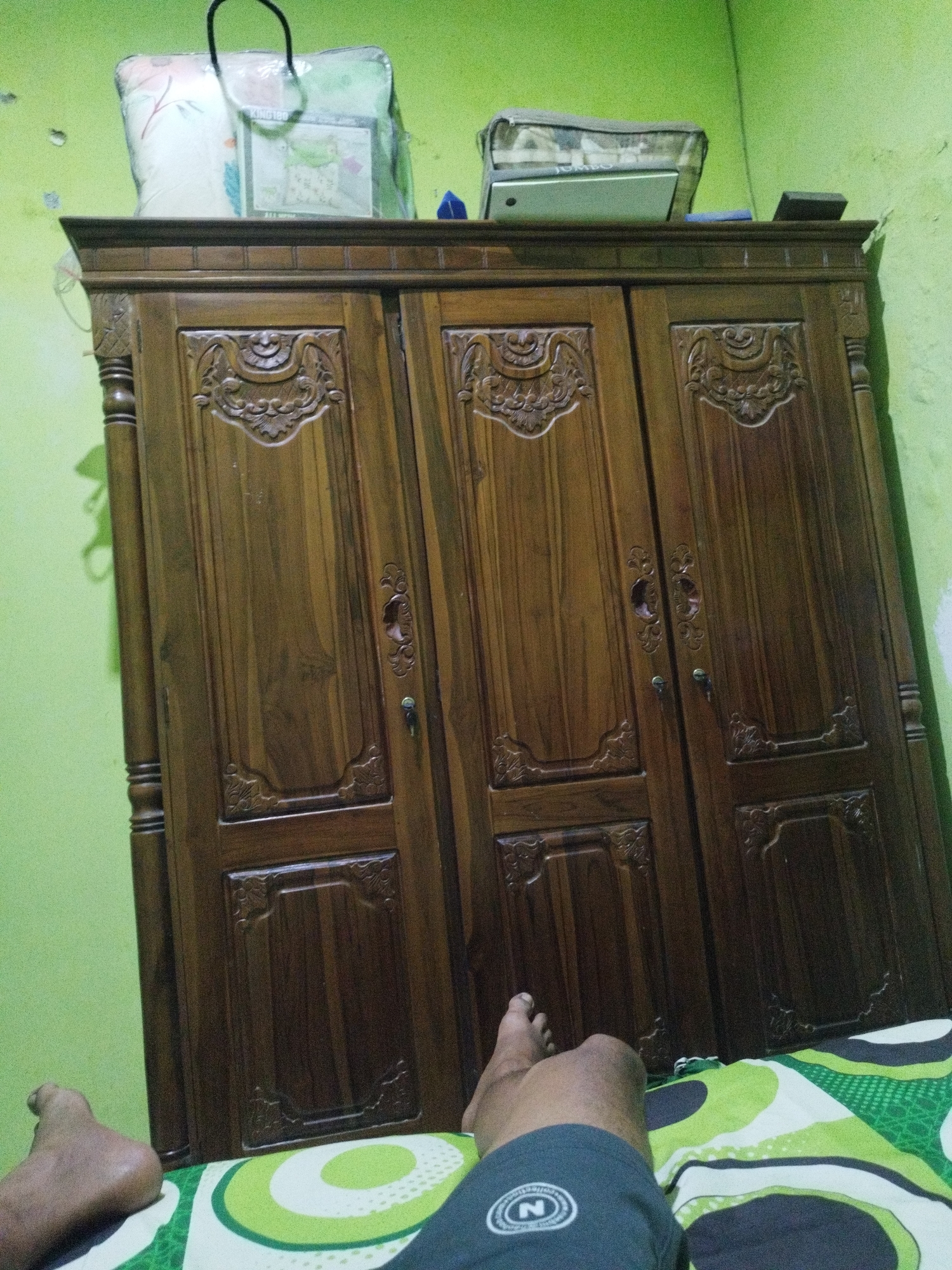 In a room with lime green walls, a large, dark wooden wardrobe with three doors dominates the scene. Each door is intricately carved with decorative patterns. On top of the wardrobe, several items are placed, including a bagged pillow with a floral design and what appears to be a closed laptop with a camouflage bag on top of it. The bottom of the image shows a person's legs and feet, lying on a bed with a green and white patterned comforter. The scene appears to be in Marengan Daya, Indonesia.