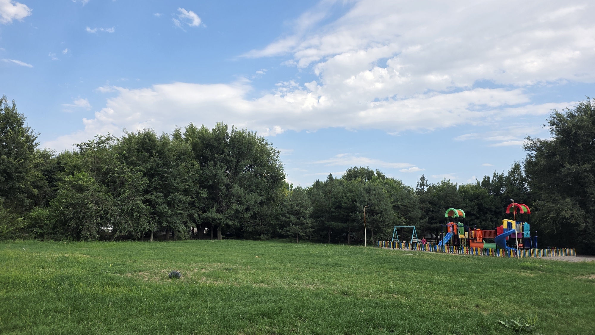 The image depicts a playground in a park setting within Tokmok City, Kyrgyzstan. The foreground is dominated by a lush green grassy field. In the middle ground to the right, a colorful playground structure is visible, featuring slides, swings, and climbing elements, enclosed by a low, multi-colored fence. The background consists of a dense line of mature trees, with a blue sky dotted with white clouds above. The lighting suggests it is daytime, likely late afternoon. There are no discernible people in the immediate vicinity of the playground. The overall scene conveys a sense of an open, recreational space.
