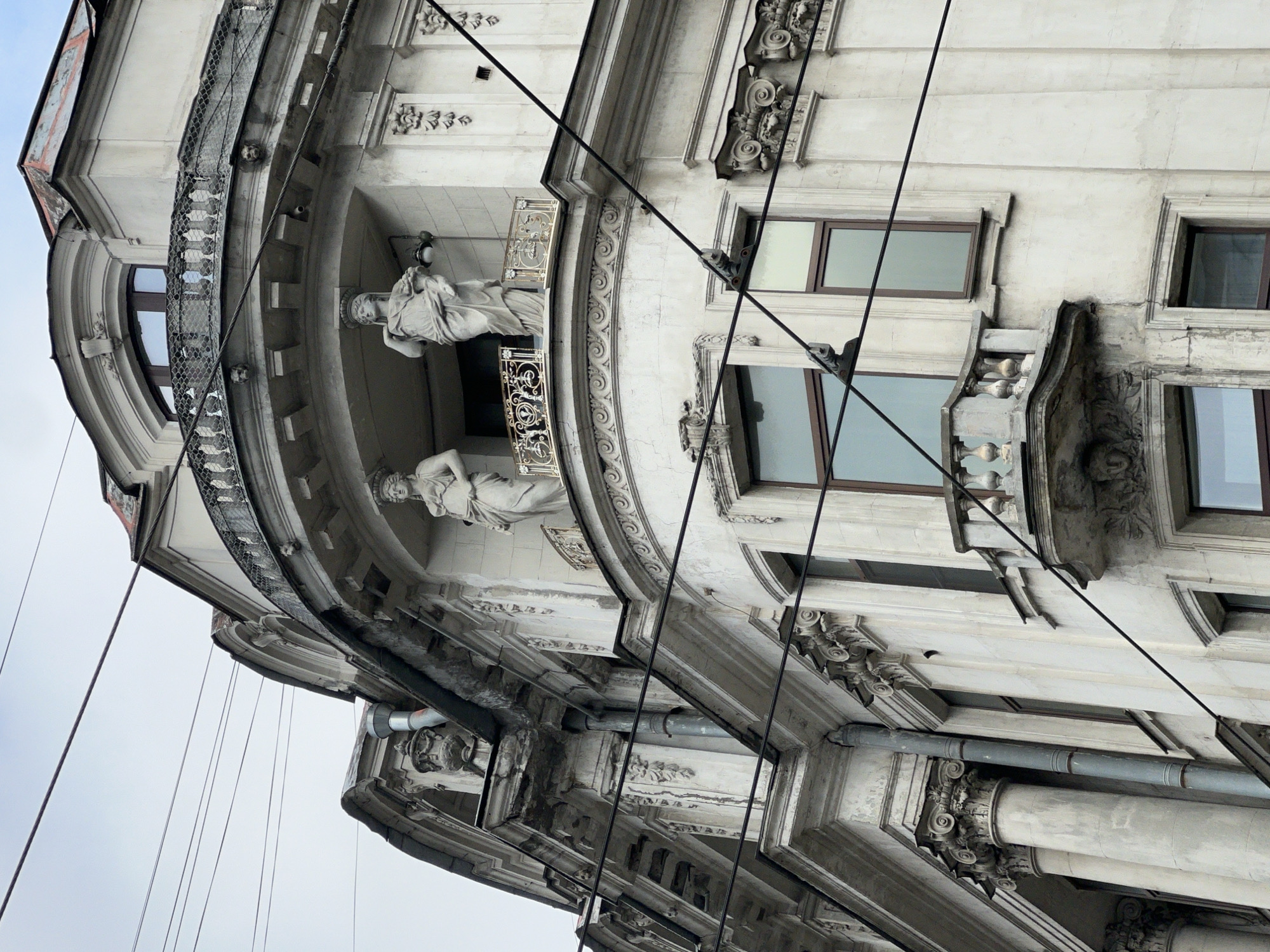 The image shows an upward view of a corner building in Saint Petersburg, Russia. The architectural style is ornate, featuring decorative molding, cornices, and details. Two classical statues stand on a rounded balcony. The building exterior is off-white or grey and seems to be made of stone or concrete. Windows are visible on the side of the building. Multiple black cables run diagonally across the scene.