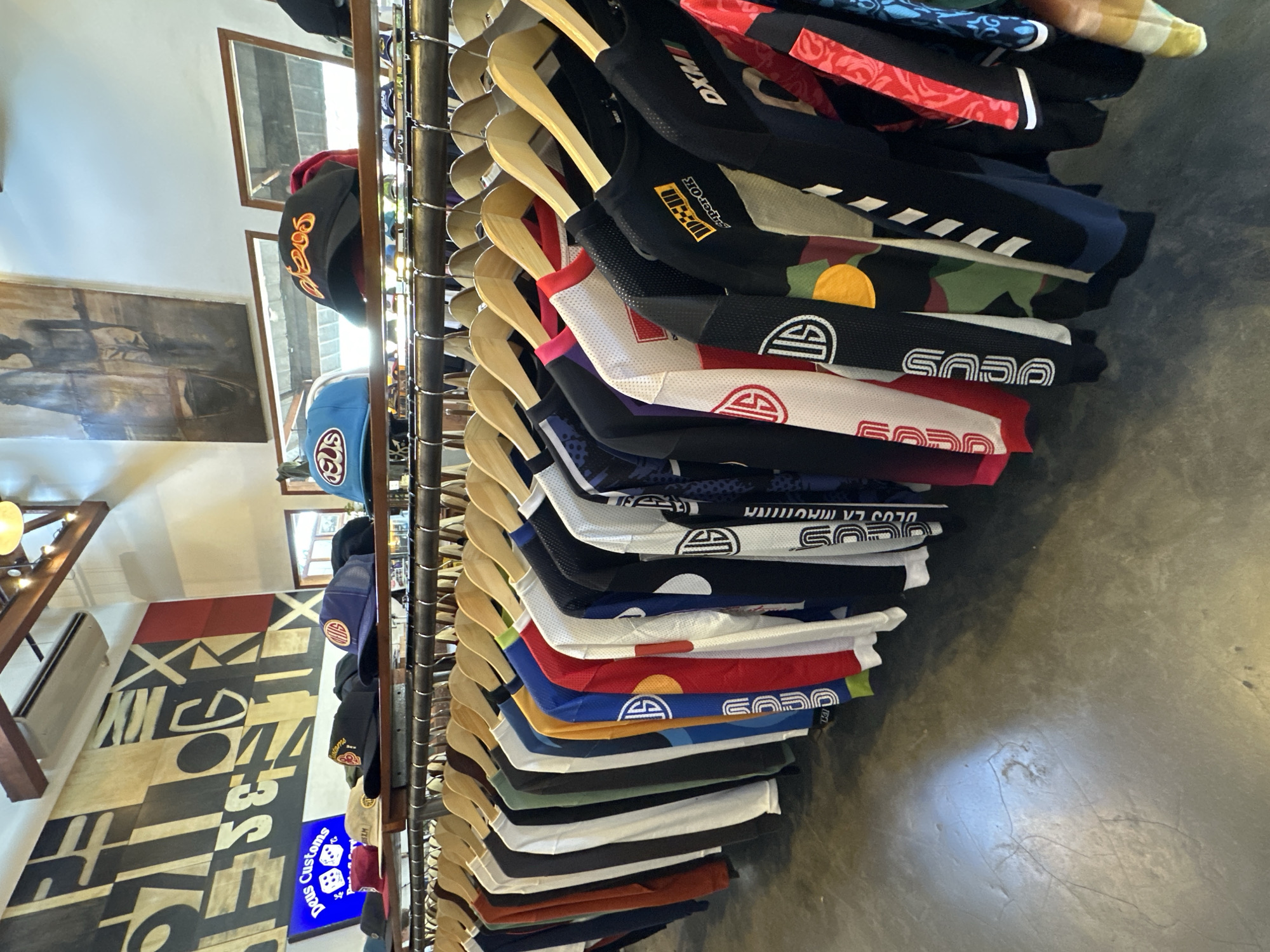 A rack of clothing hangs vertically in a shop. The clothes appear to be jerseys or shirts of various colors and patterns. The word "SODO" is visible on several of the garments. Wooden hangers support the clothes. In the background, hats hang on a separate rack, and the wall is decorated with abstract designs. A sign that reads "Deus Customs" is also visible. The location is North Kuta, Indonesia.