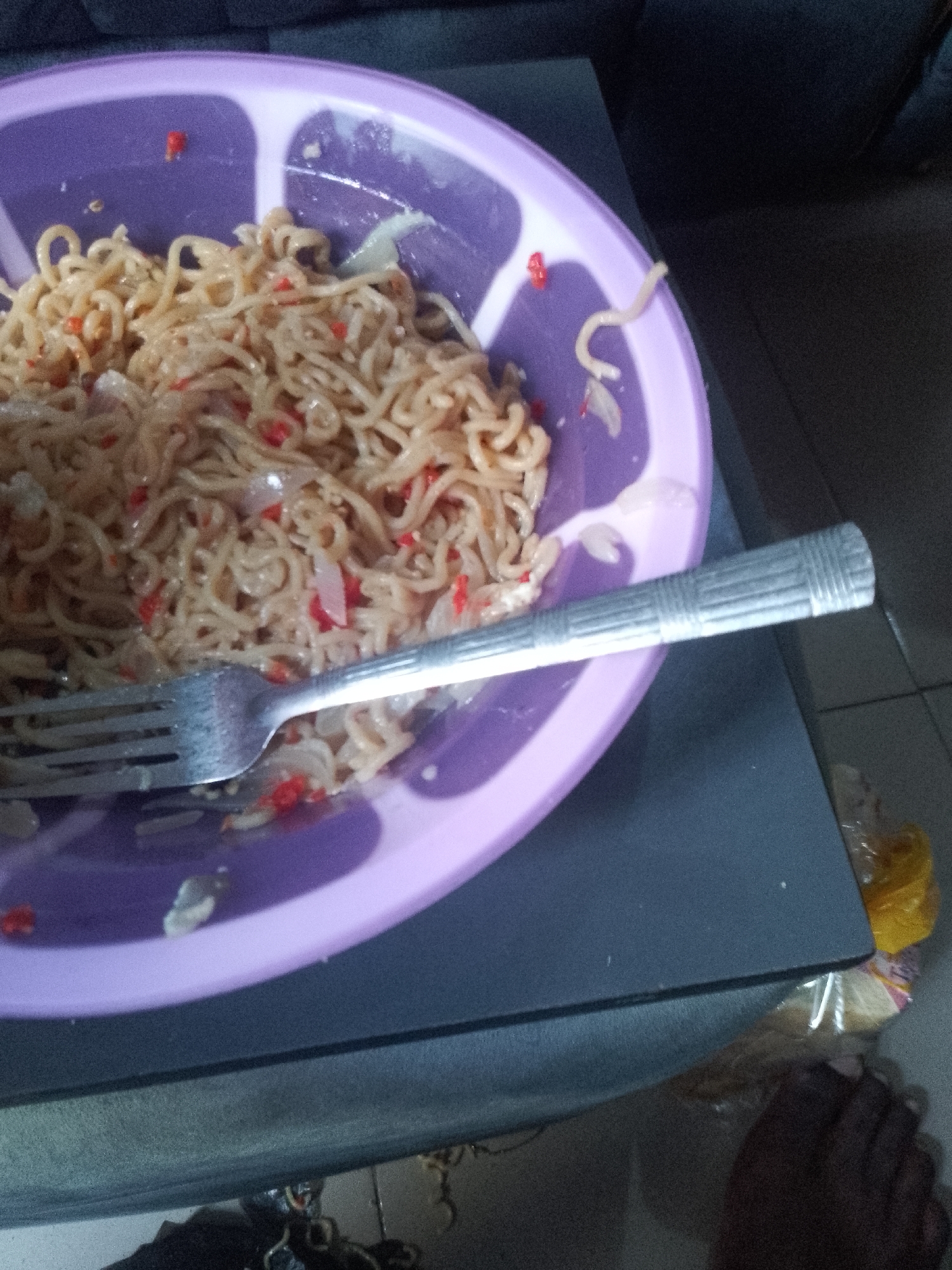 An overhead shot shows a purple bowl filled with instant noodles, garnished with chopped red peppers and onions. A silver fork rests on the rim of the bowl, with its tines submerged in the noodles. The bowl is placed on a dark gray table, with a dark sofa visible in the background. A person's dark-skinned foot and ankle are visible in the lower right corner of the frame, with a plastic bag containing food items nearby. The scene appears to be indoors.