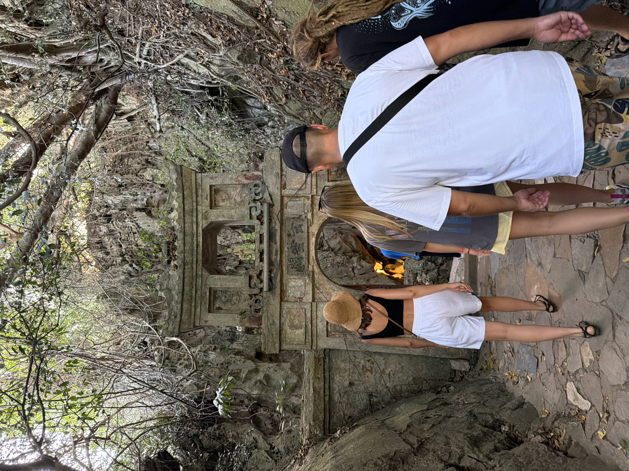 This image captures a group of four individuals walking towards a large, ornate stone archway that leads into a cave or grotto. The archway appears to be a temple or shrine, with intricate carvings and statues visible at its peak. Lush vegetation and gnarled tree roots frame the entrance, suggesting a natural, somewhat overgrown setting. The ground is paved with irregular stones.
The individuals are walking away from the camera, towards the archway. The person in the foreground, wearing a white t-shirt and camouflage shorts, has a black strap across their chest. To their left, a person in a black top and white shorts wears a straw hat. To the right of the person in the white t-shirt, another individual with long, blonde hair wears a grey t-shirt and yellow shorts. On the far right, a person with dreadlocks wears a black t-shirt. The scene conveys a sense of exploration and discovery, likely in a historical or religious site.
The location is Marble Mountains (Ngũ Hành Sơn) in Đà Nẵng, Vietnam. Specifically, this appears to be the entrance to the Huyen Khong cave (Hang Huyen Khong) or a similar cave within the Marble Mountains, known for its Buddhist shrines and natural caves that have been adapted for religious purposes. The distinctive rock formations, dense foliage, and the architectural style of the archway are characteristic of this area.