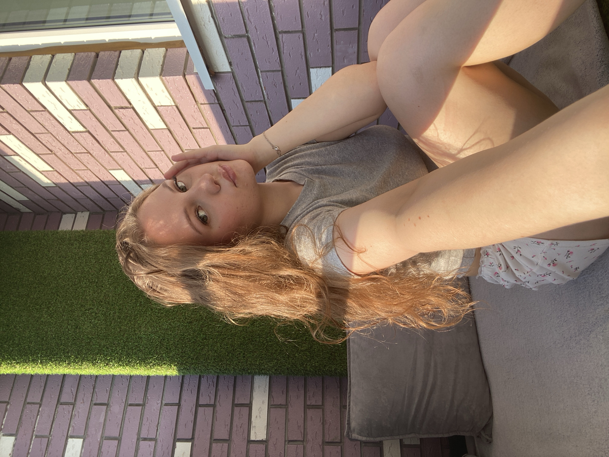 A young woman with long, auburn hair lies on a grey sofa on a patio. She wears a grey t-shirt and floral shorts. The patio has artificial grass and brick flooring, with light-colored bricks interspersed with darker ones. The location is possibly a balcony, but it is impossible to confirm that this image was taken in the city of Izhevsk, Russia, or not.