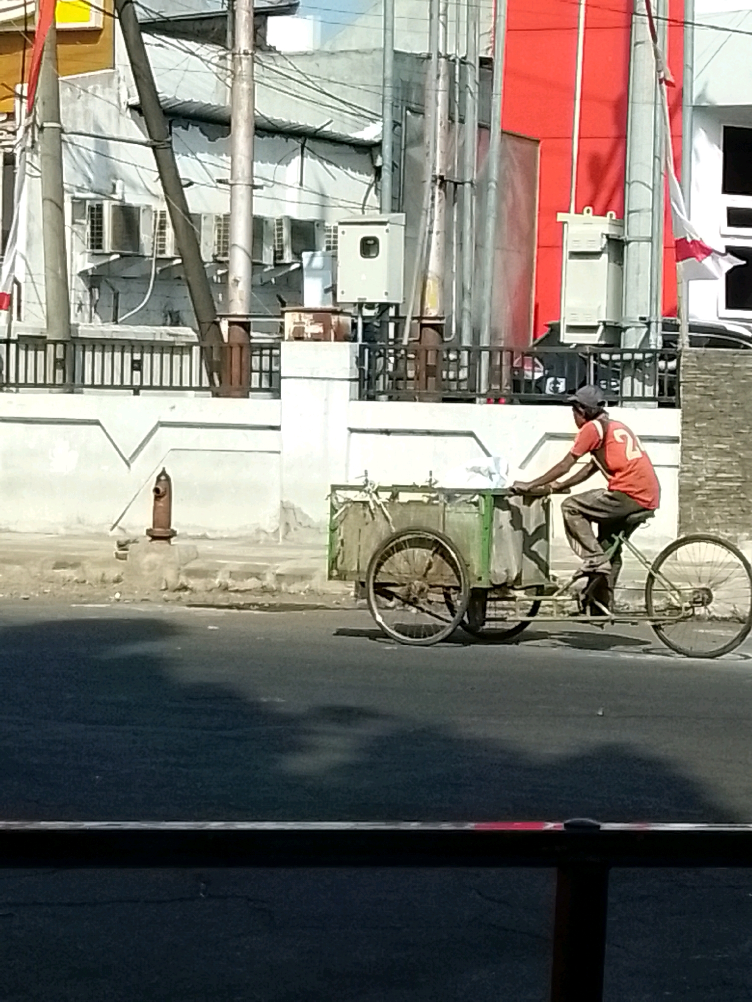 A man in an orange shirt with the number 24 on the back is riding a three-wheeled bicycle with a cargo box in front. The bicycle is on a paved road, casting a shadow. Behind the road is a sidewalk with a fire hydrant and a white wall with a decorative pattern. In the background, there are buildings, including one with a red facade and a white one with air conditioning units. Electrical wires and poles are visible overhead. The scene appears to be in a city, possibly Jember, Indonesia, given the architectural style and the presence of flags. The man is wearing a dark cap and dark pants. The cargo box on the bicycle is green and appears to contain white plastic bags.